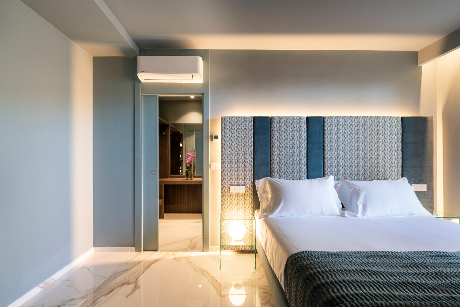 Modern hotel bedroom featuring a stylish bed with a patterned headboard, marble flooring, and soft lighting. The room includes an air conditioning unit and an open doorway leading to a bathroom area. Ideal for travelers seeking comfort and elegance. | Only on SopranoVillas