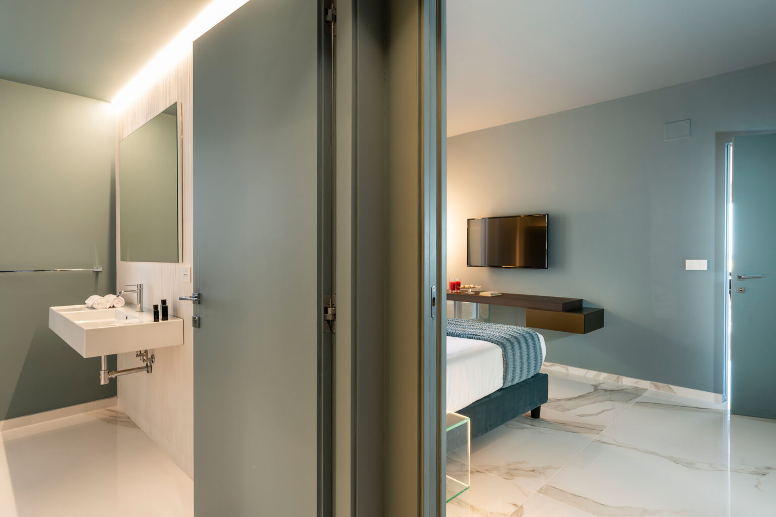 Modern hotel room with an attached bathroom, featuring a minimalist design, soft lighting, a comfortable bed, and a flat-screen TV on the wall. The bathroom includes a stylish sink and contemporary fixtures. | Only on SopranoVillas
