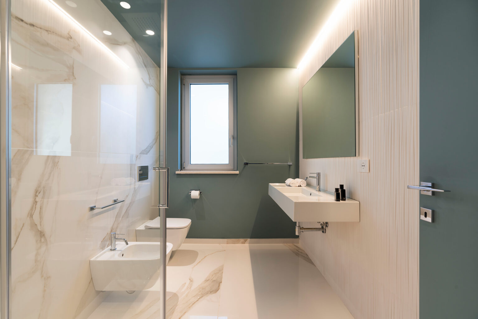 Modern bathroom design featuring a glass shower, bidet, and wall-mounted sink. The walls are a calming green with elegant marble accents, complemented by natural light from a window. Ideal for showcasing contemporary interior aesthetics. | Only on SopranoVillas