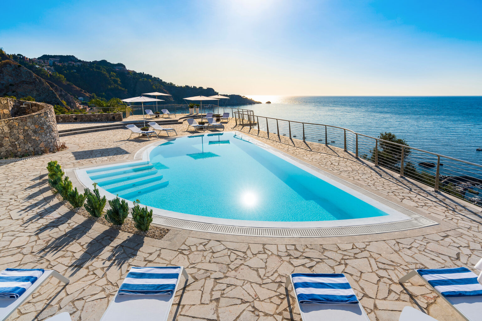 Stunning ocean view from a luxury villa with a modern swimming pool, surrounded by sun loungers and lush greenery, under a clear blue sky at sunset. | Only on SopranoVillas