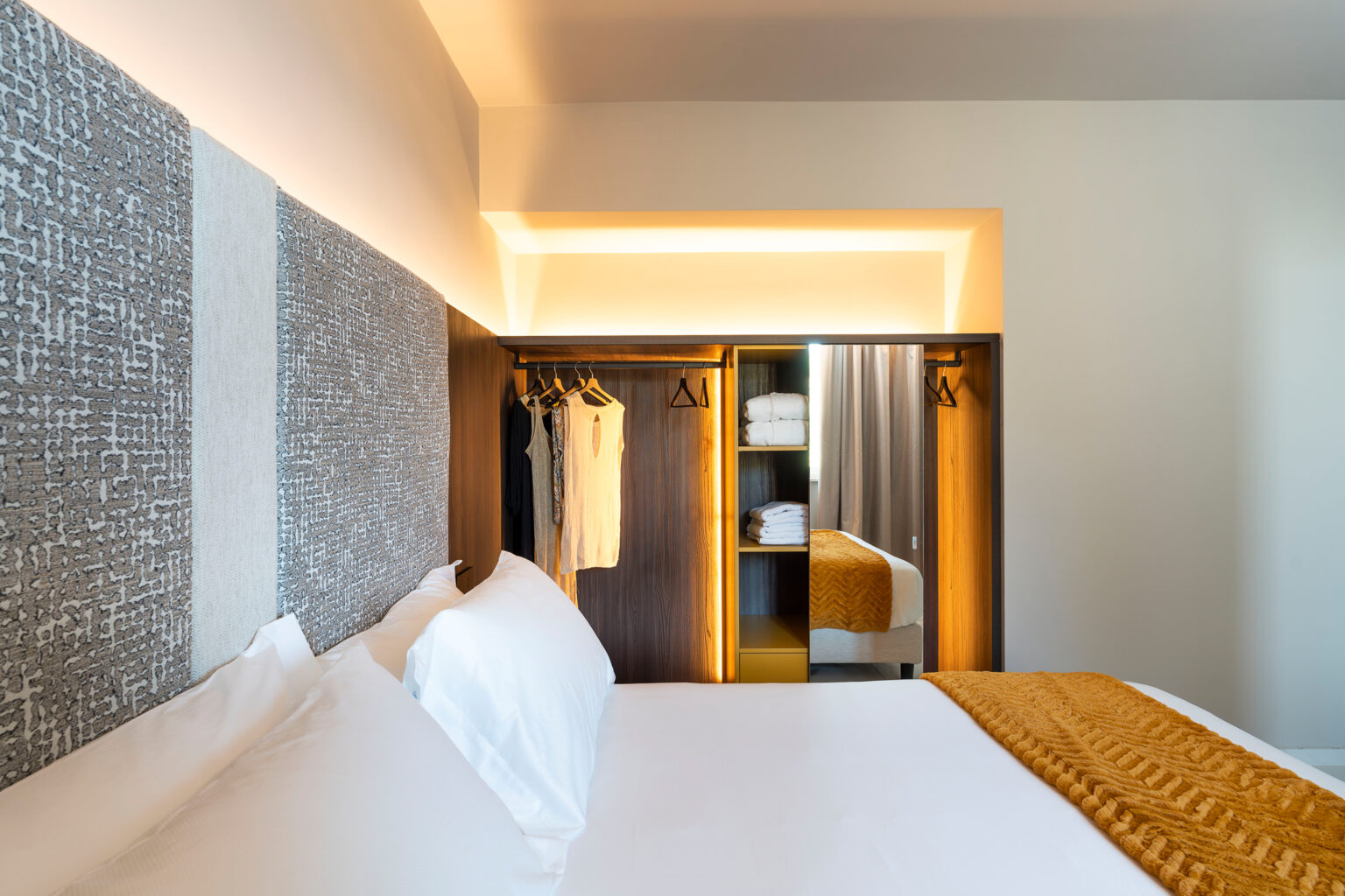 Modern hotel room featuring a comfortable bed with white linens, a stylish accent wall, and a well-organized wardrobe displaying clothing and towels. Soft lighting enhances the cozy atmosphere. | Only on SopranoVillas