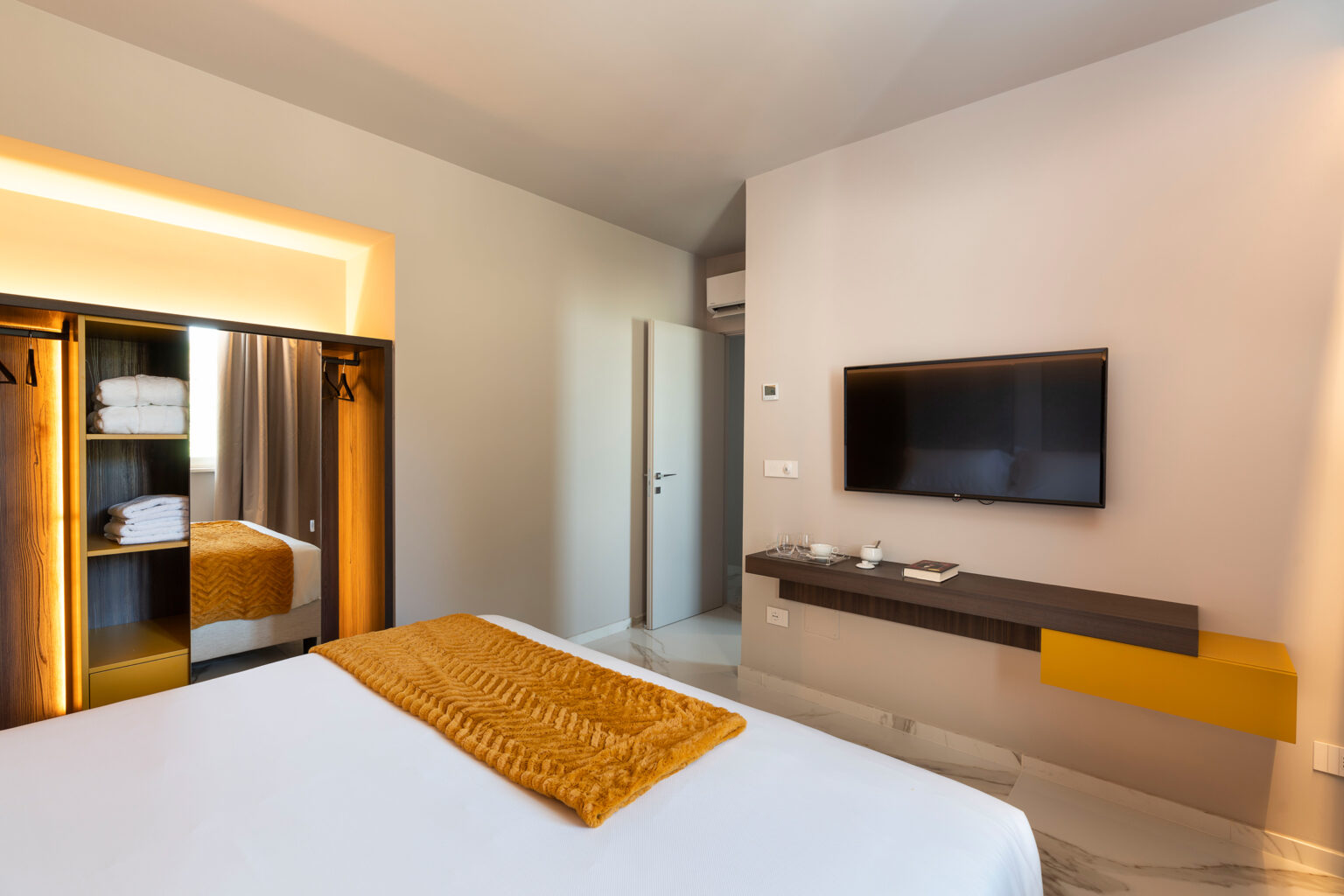 Modern hotel room featuring a comfortable bed with an orange throw blanket, a flat-screen TV mounted on the wall, and a stylish wardrobe. The room is well-lit with contemporary decor and includes a door leading to a bathroom. Ideal for travelers seeking a relaxing stay. | Only on SopranoVillas