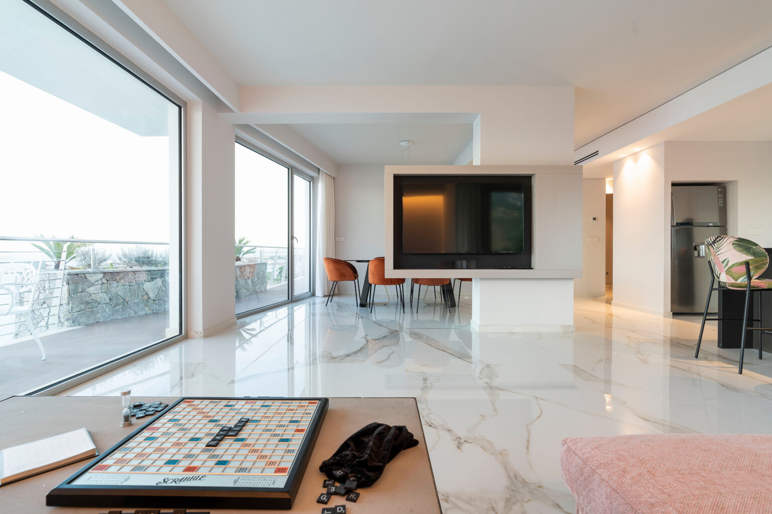 Modern living room featuring a marble floor, a Scrabble board game on a coffee table, and large windows with a view of the balcony. The space includes a sleek TV unit and stylish dining area with orange chairs, creating an inviting and contemporary atmosphere. | Only on SopranoVillas