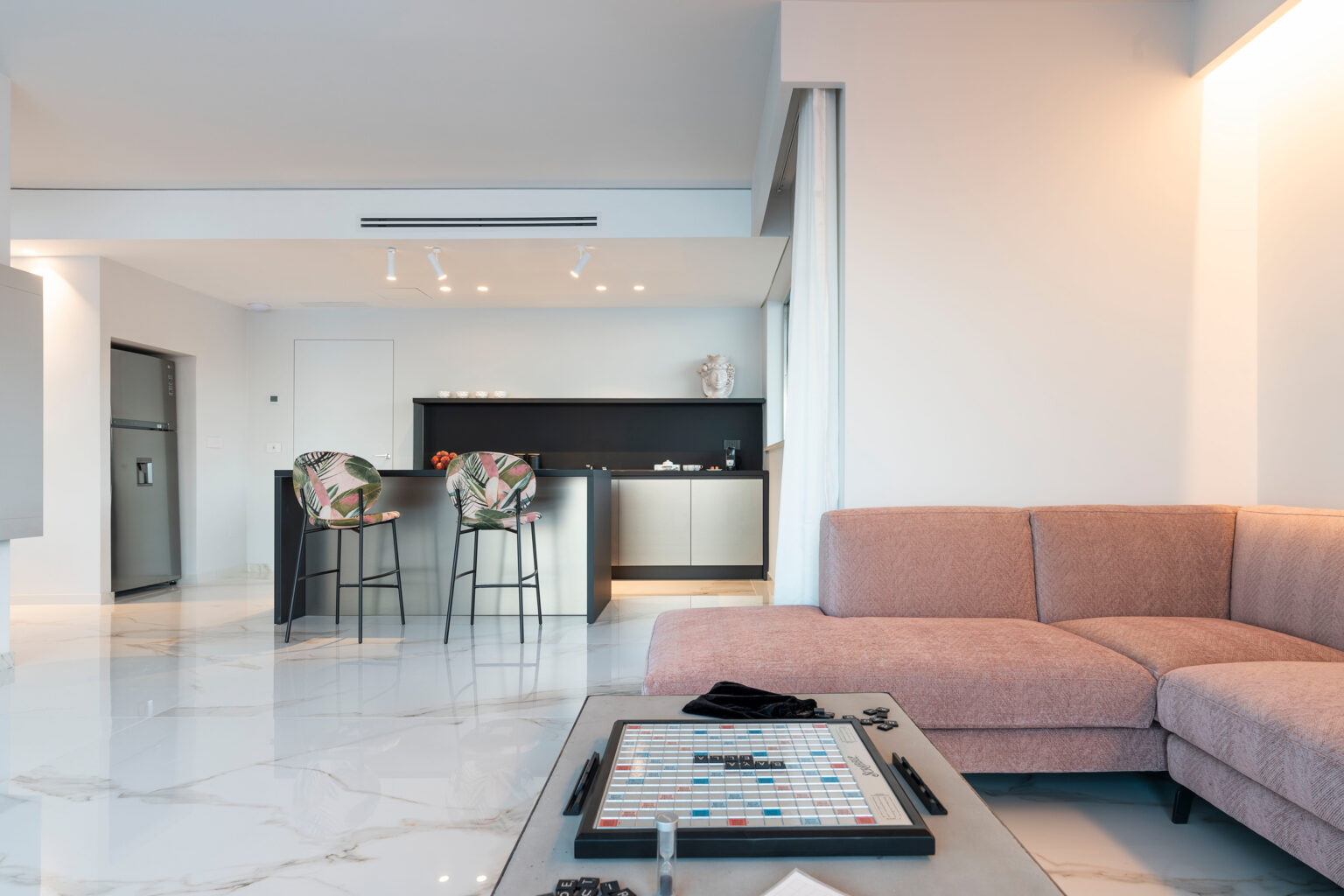 Modern living room with a pink sectional sofa, a coffee table, and a stylish kitchen in the background featuring bar stools and sleek appliances. Bright, airy atmosphere with marble flooring and contemporary decor. | Only on SopranoVillas