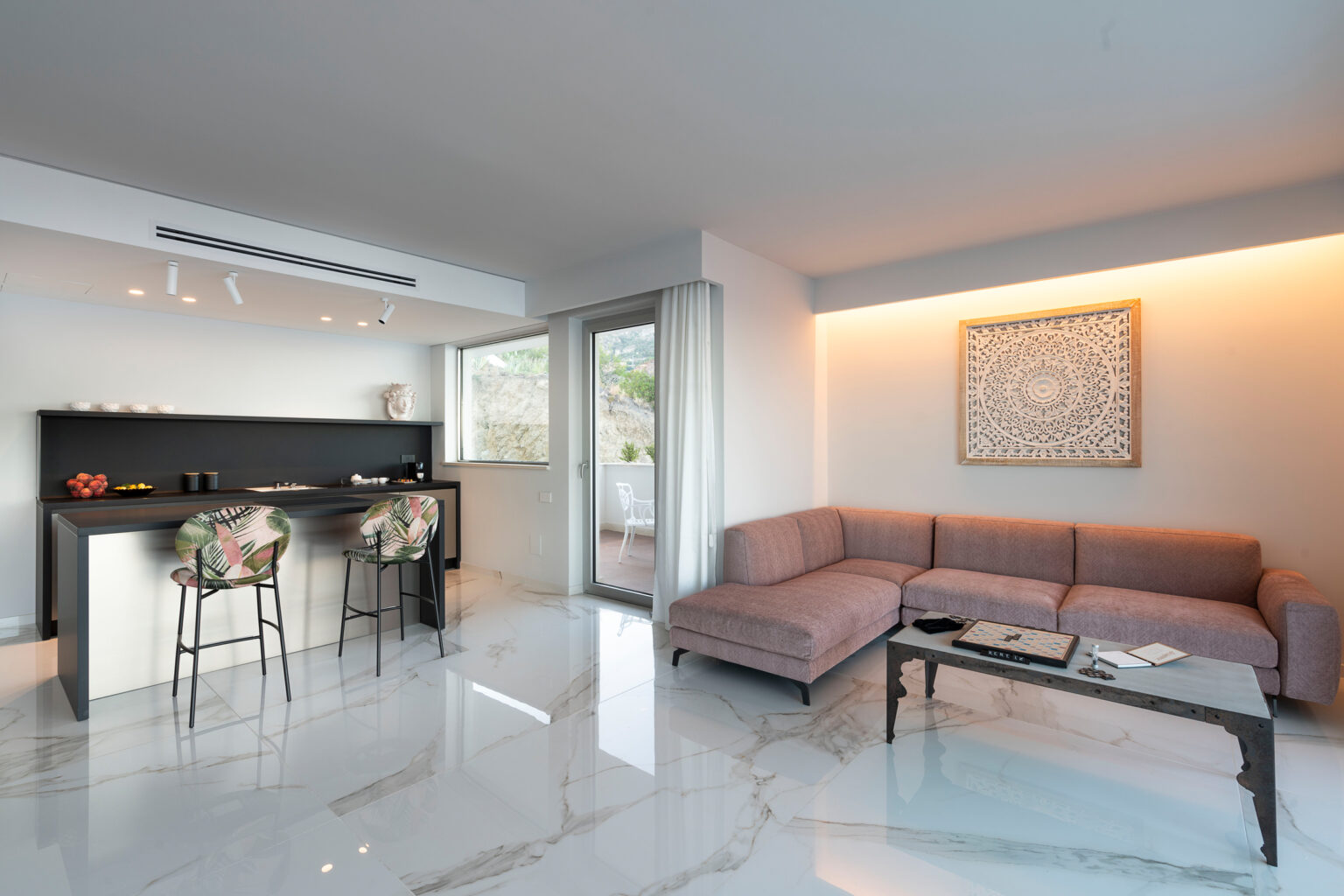 Modern living room with a sleek design featuring a comfortable sectional sofa, a minimalist coffee table, and a stylish kitchen bar with stools. Bright, airy space enhanced by natural light and elegant marble flooring. | Only on SopranoVillas
