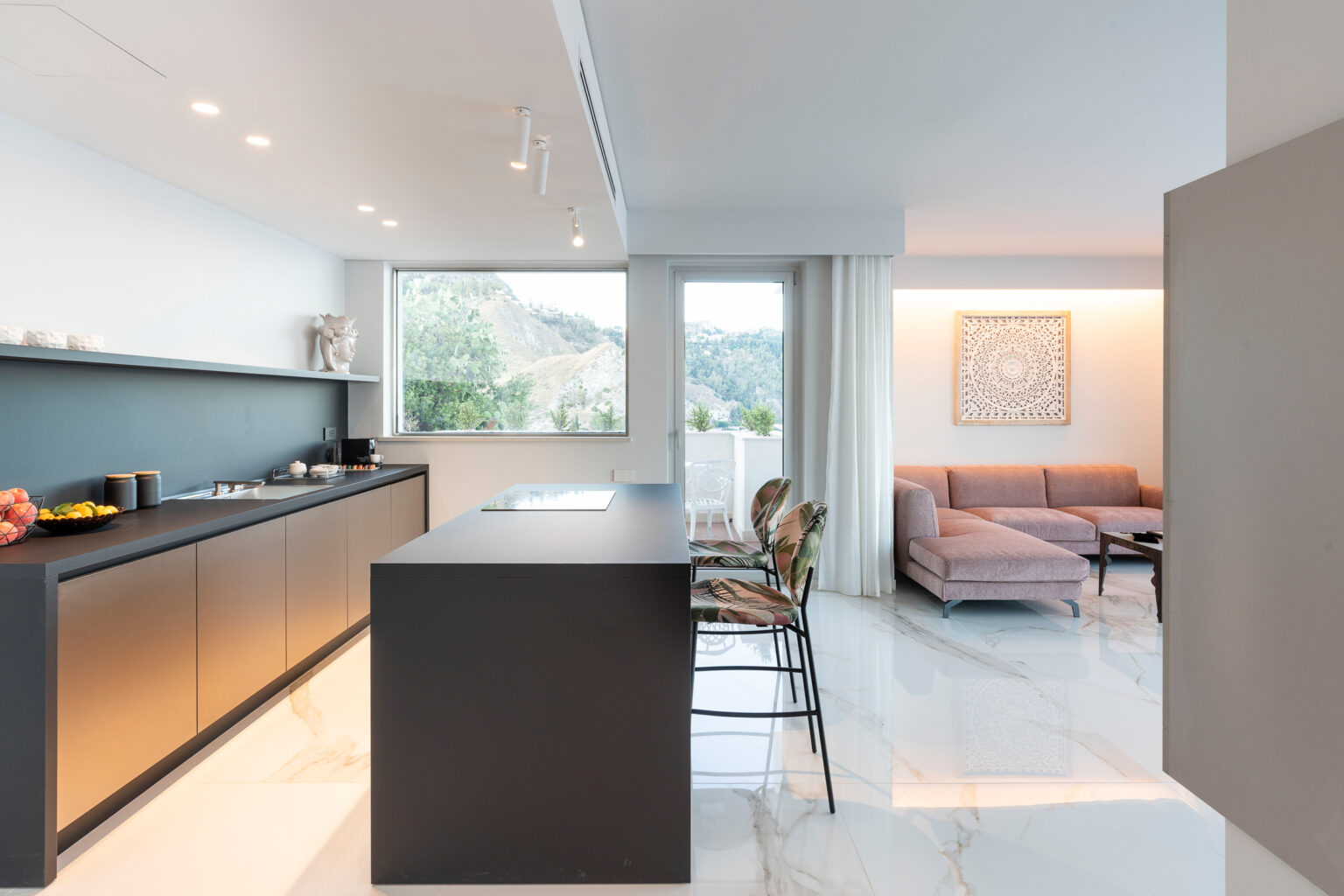 Modern kitchen and living area featuring sleek cabinetry, a spacious island, and natural light from large windows. The design includes minimalist decor with a cozy seating area in the background, showcasing a blend of functionality and style. | Only on SopranoVillas