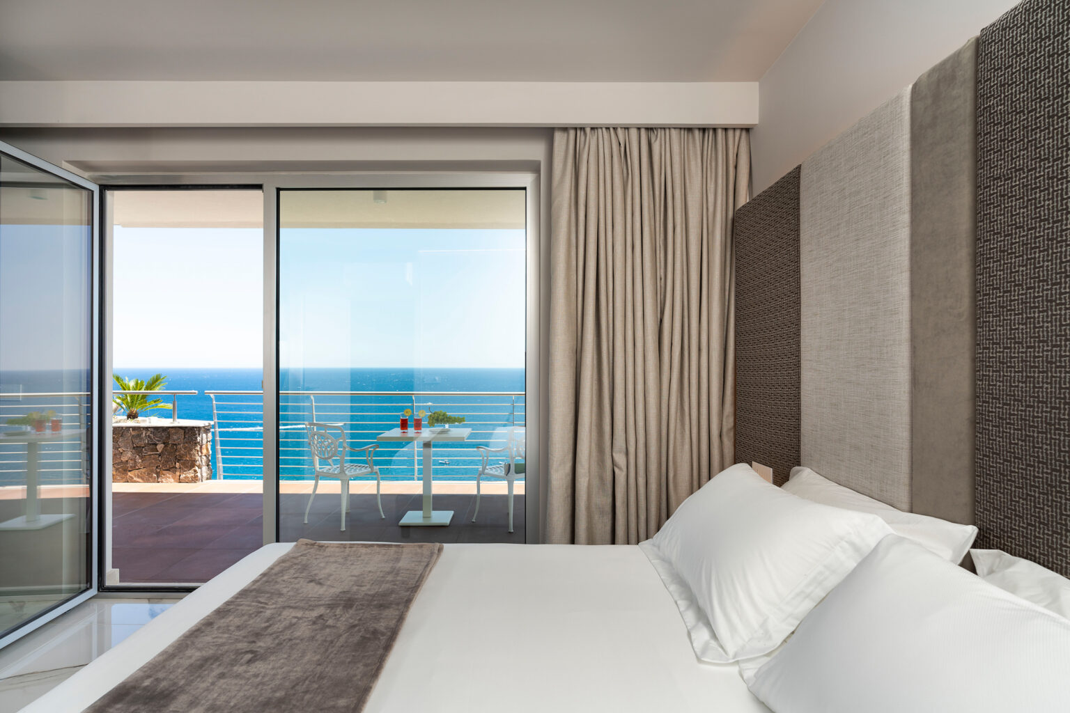 Modern hotel bedroom with a king-size bed, featuring large glass doors that open to a balcony with ocean views. Elegant decor includes neutral tones and soft furnishings, creating a serene atmosphere. Ideal for luxury travelers seeking a coastal getaway. | Only on SopranoVillas