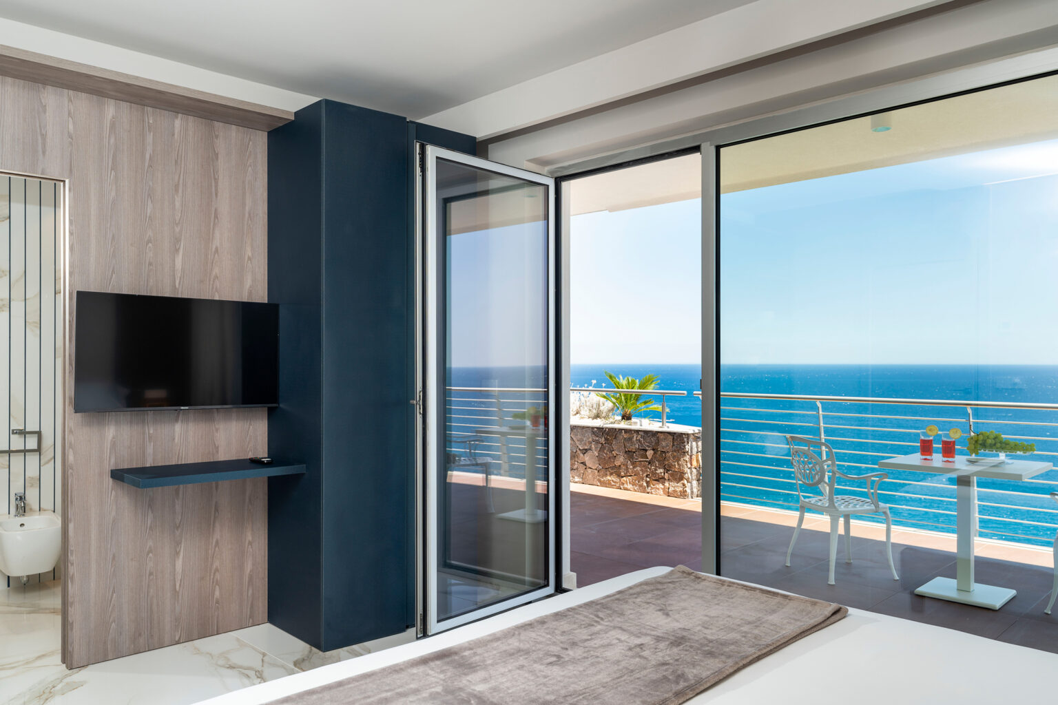 Modern hotel room with a stunning ocean view, featuring a comfortable bed, flat-screen TV, and large sliding glass doors leading to a balcony with outdoor seating. | Only on SopranoVillas