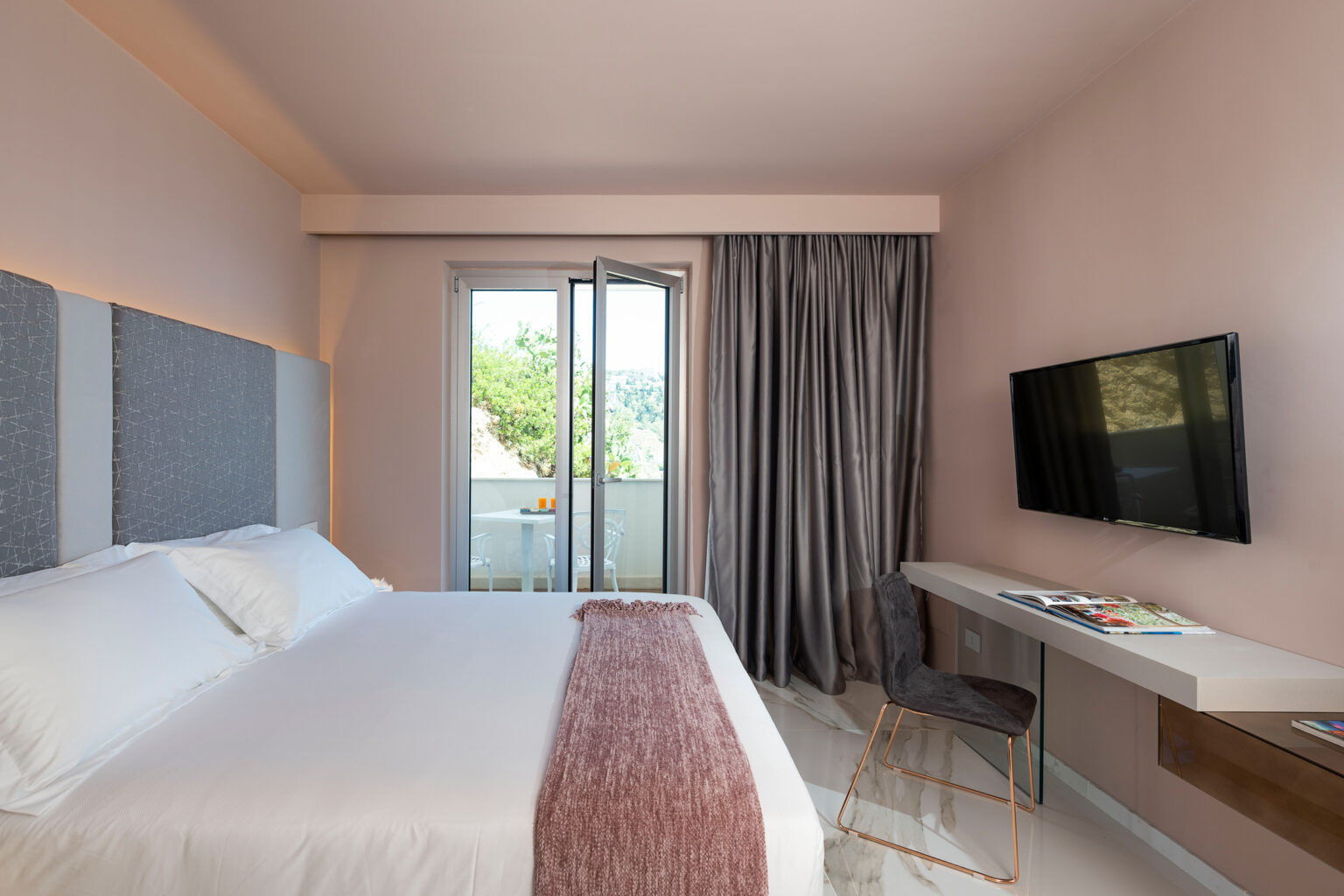Modern hotel room featuring a queen-sized bed, stylish decor, a flat-screen TV, and a small workspace. Natural light streams in from the balcony doors, complemented by elegant curtains and a minimalist design. Perfect for a comfortable stay. | Only on SopranoVillas