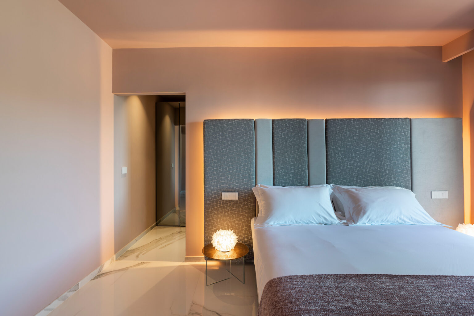 Modern bedroom interior featuring a neatly made bed with gray upholstered headboard, soft lighting, and a minimalist design. The room has a warm color palette with a view of a bathroom entrance. Perfect for showcasing contemporary home decor and comfort. | Only on SopranoVillas