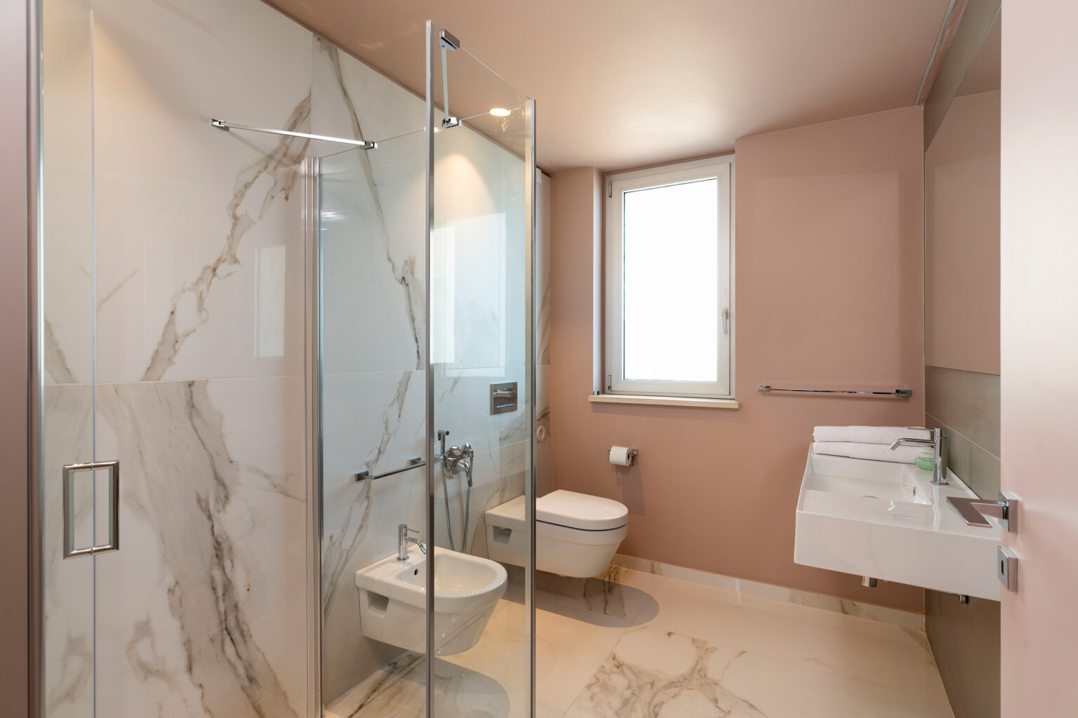 Modern bathroom featuring a glass shower enclosure, marble walls, a wall-mounted sink, and a bidet, with natural light coming through a window. | Only on SopranoVillas