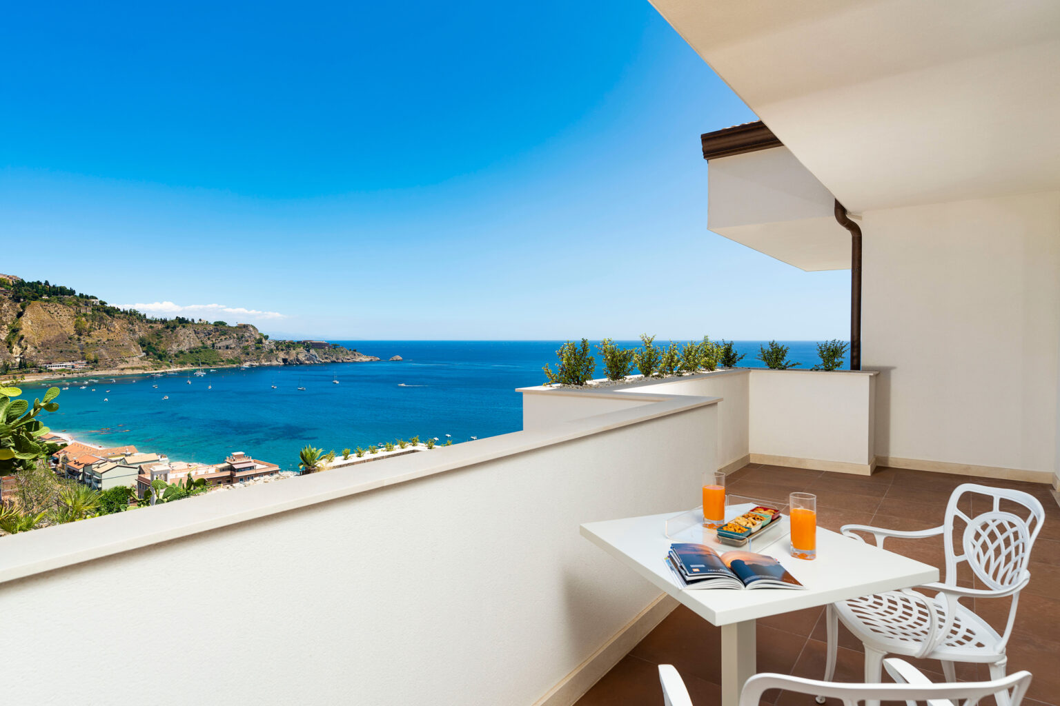 Bright balcony with a scenic ocean view, featuring a small white table with two orange drinks and a magazine, overlooking a coastal landscape. | Only on SopranoVillas