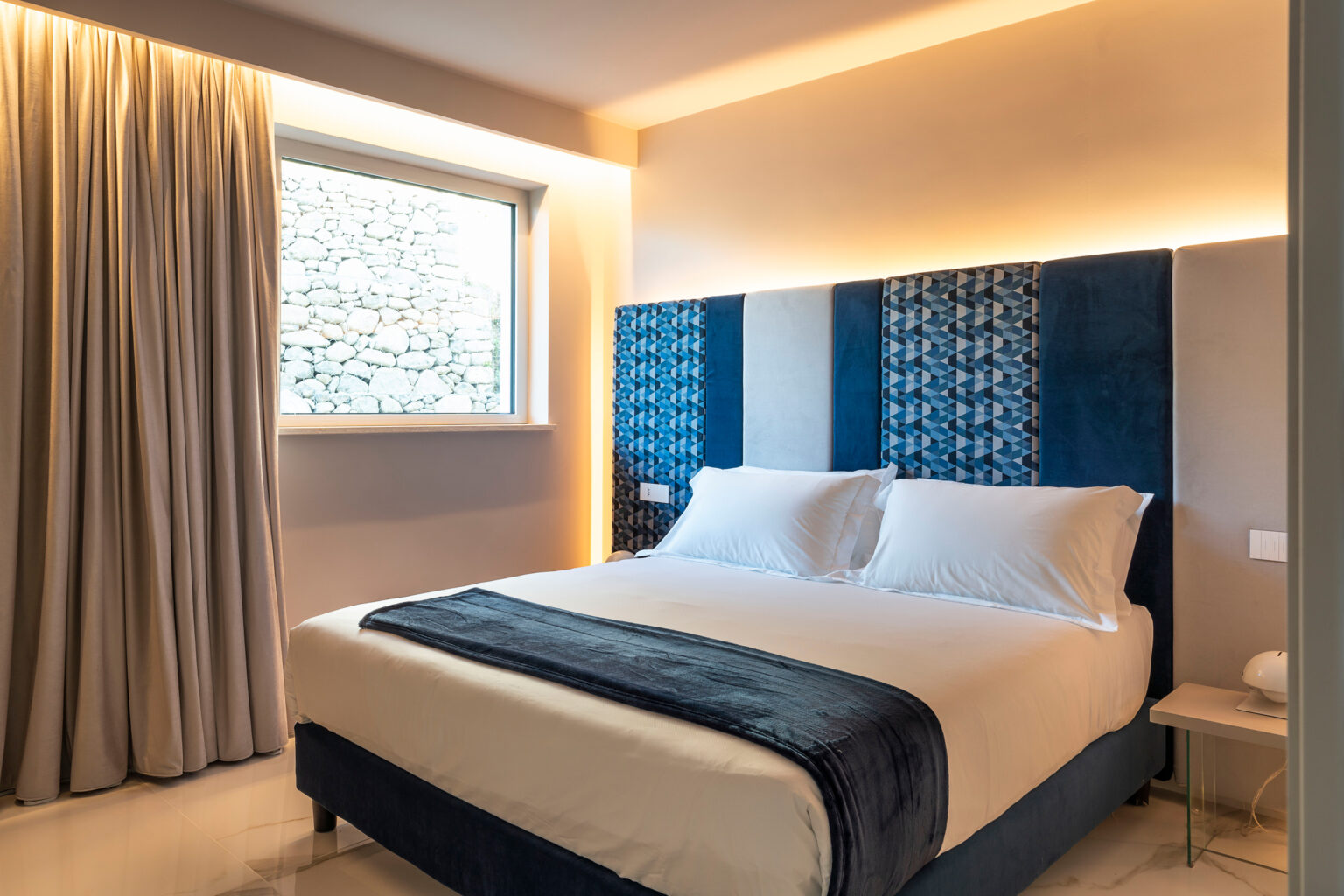 Modern hotel bedroom featuring a large double bed with white linens, stylish blue headboard, soft lighting, and elegant curtains. The room has a minimalist design with a cozy ambiance, ideal for relaxation. | Only on SopranoVillas
