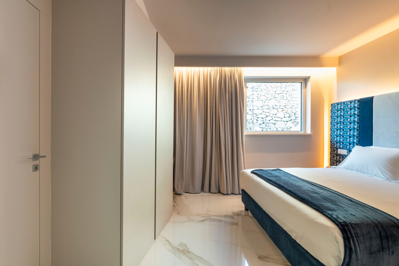 Modern hotel bedroom featuring a comfortable bed, stylish blue headboard, and large window with sheer curtains, complemented by sleek marble flooring and minimalist decor. | Only on SopranoVillas