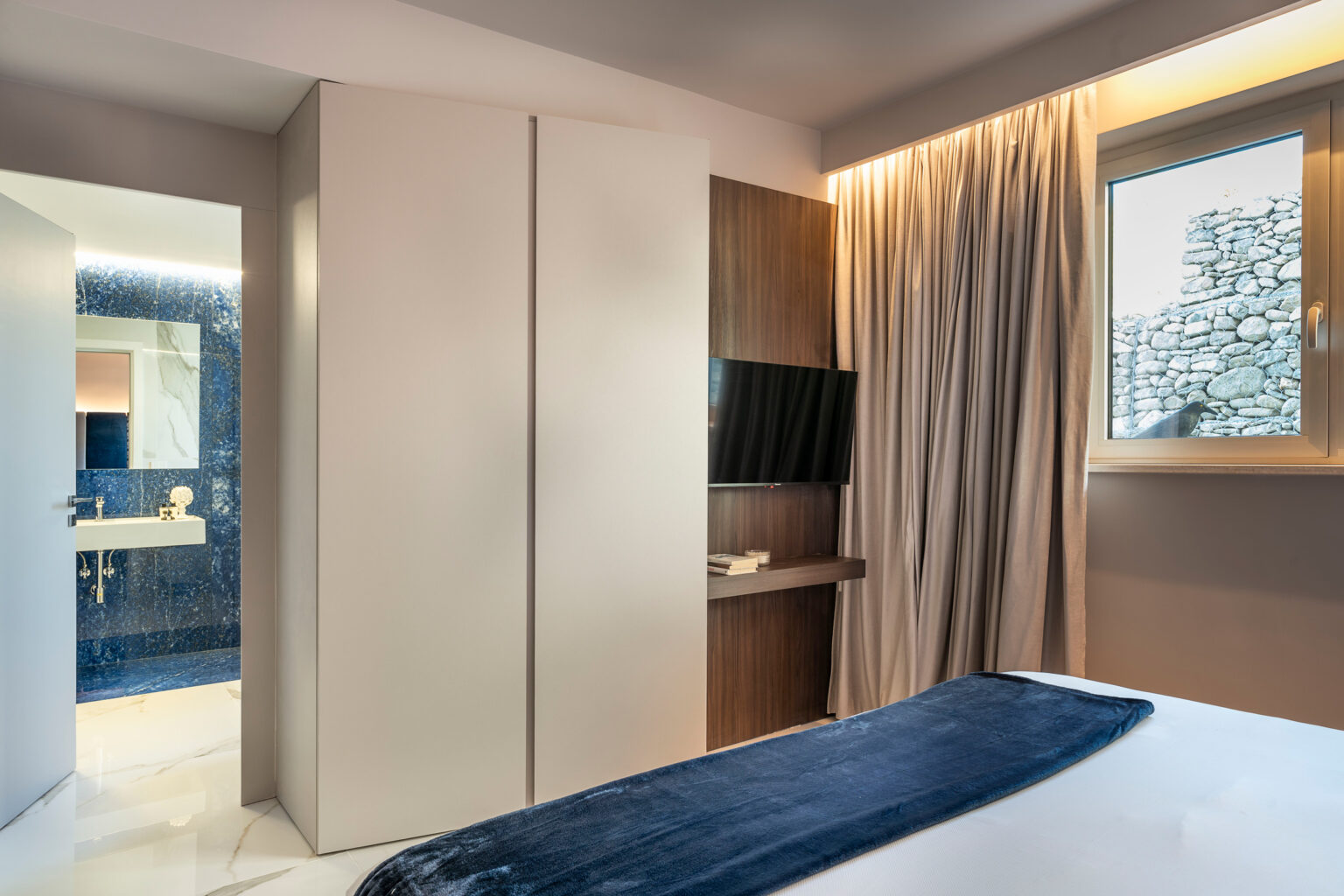 Modern hotel room featuring a sleek wardrobe, a wall-mounted TV, and large windows with natural light. The decor includes neutral tones and soft textures, creating a cozy atmosphere. A bathroom can be seen through the open door, showcasing contemporary design elements. | Only on SopranoVillas