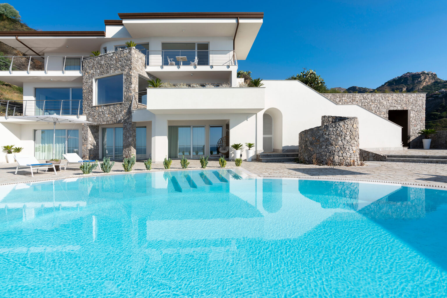 Luxurious modern villa with a large swimming pool, surrounded by stone and greenery, set against a clear blue sky. | Only on SopranoVillas