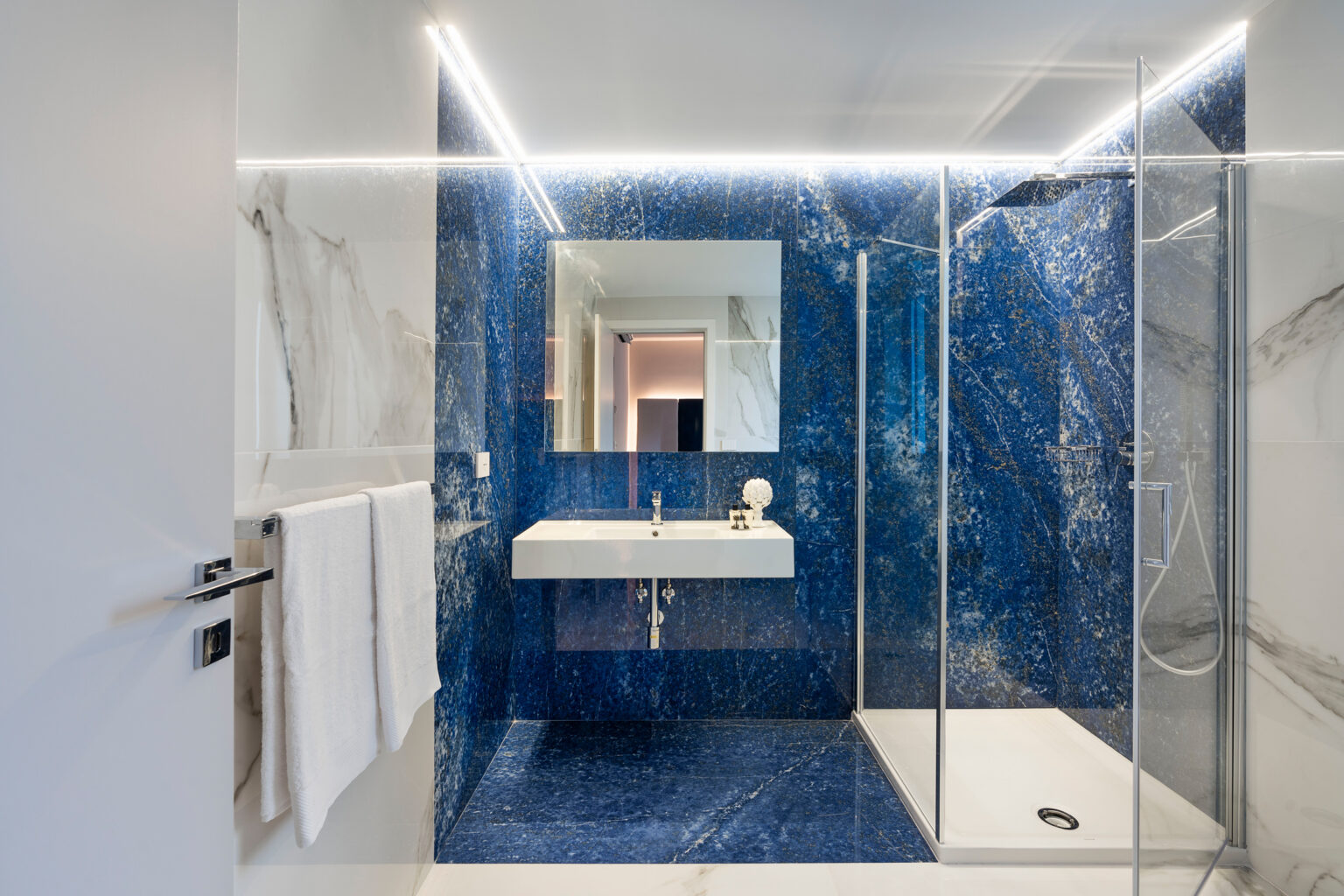 Modern bathroom featuring striking blue marble walls, a sleek white sink, and a glass shower enclosure, illuminated by LED lighting. | Only on SopranoVillas