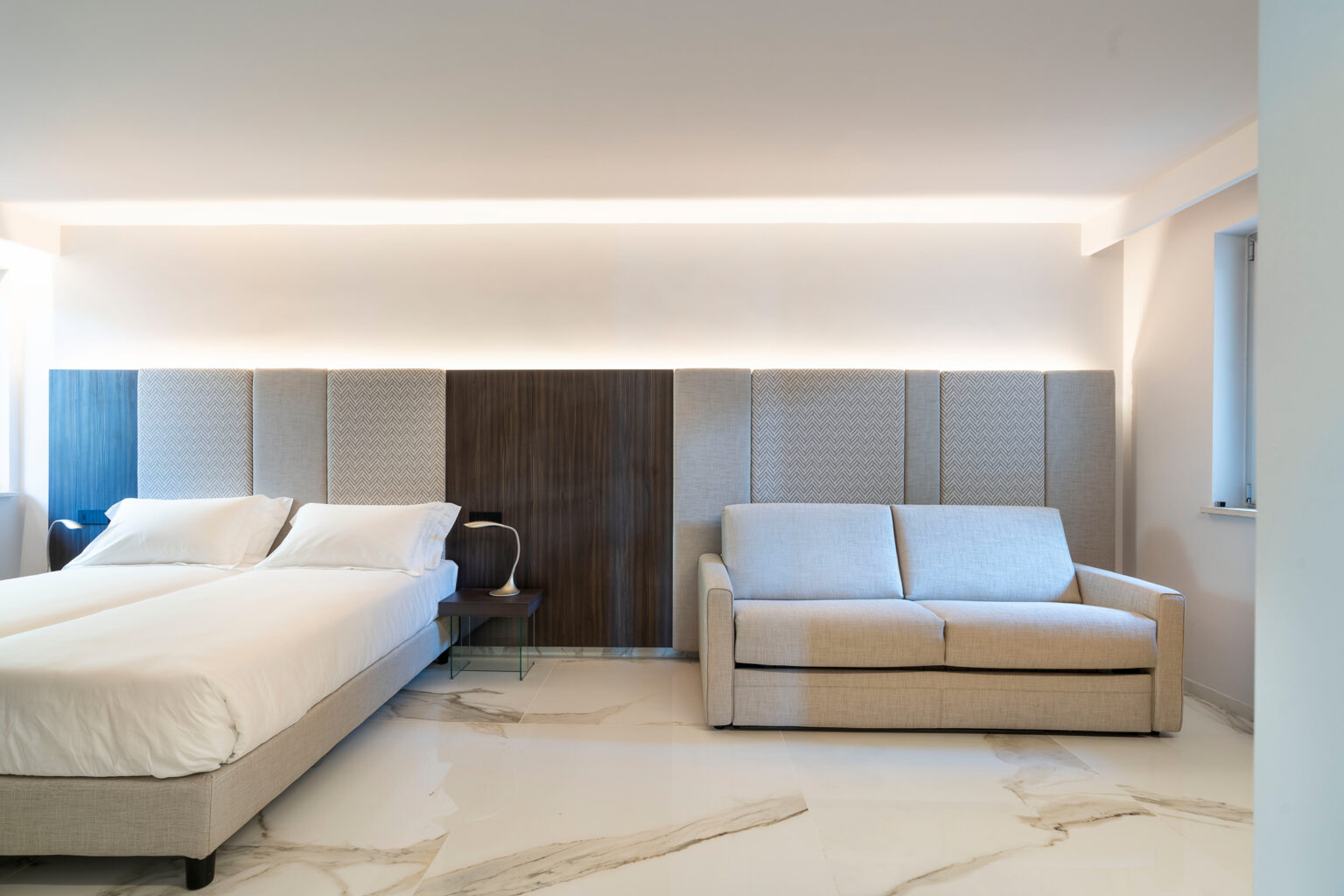 Modern bedroom interior featuring a king-size bed with crisp white linens, a minimalist design, and a comfortable sofa. Soft ambient lighting enhances the spacious atmosphere, complemented by elegant marble flooring. | Only on SopranoVillas