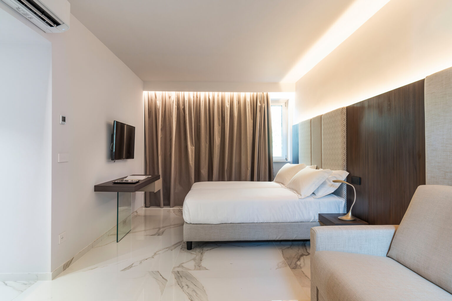 Modern hotel room featuring a queen bed, minimalist decor, a flat-screen TV, and elegant lighting. The space includes a comfortable seating area and large windows with curtains, showcasing a stylish and inviting atmosphere. | Only on SopranoVillas