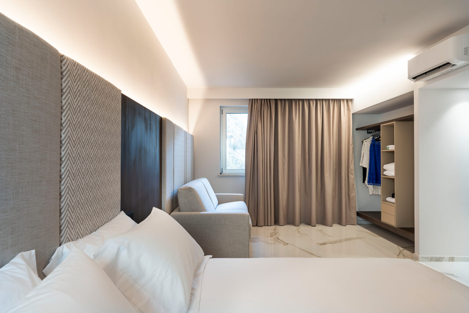 Modern hotel room featuring a comfortable bed, stylish gray curtains, and a cozy seating area. The room is well-lit with contemporary decor and includes a wardrobe with neatly arranged clothing. | Only on SopranoVillas