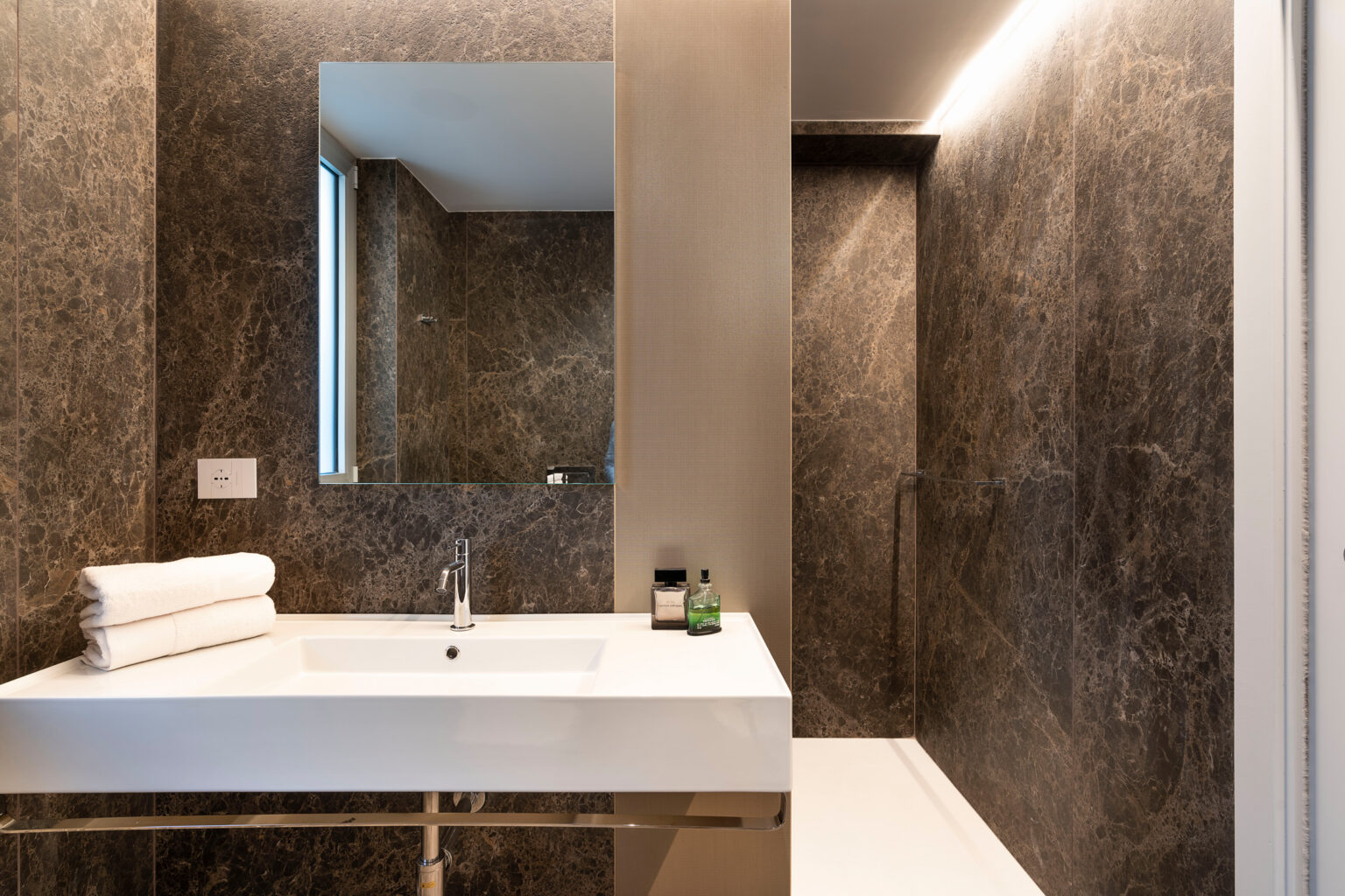 Modern bathroom featuring a sleek white sink, neatly stacked towels, and elegant dark stone walls. The space includes a large mirror and a glass shower enclosure, creating a luxurious and contemporary atmosphere. Ideal for home renovation inspiration. | Only on SopranoVillas