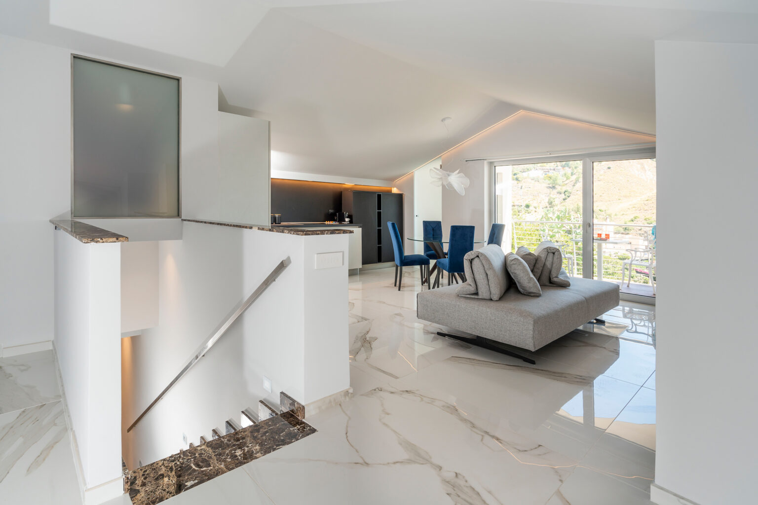 Modern living room interior featuring a stylish gray sofa with pillows, a glass dining table surrounded by blue chairs, and a sleek kitchen area. The space is illuminated by natural light from large windows, showcasing a scenic view. Elegant marble flooring and contemporary decor elements enhance the aesthetic appeal. | Only on SopranoVillas