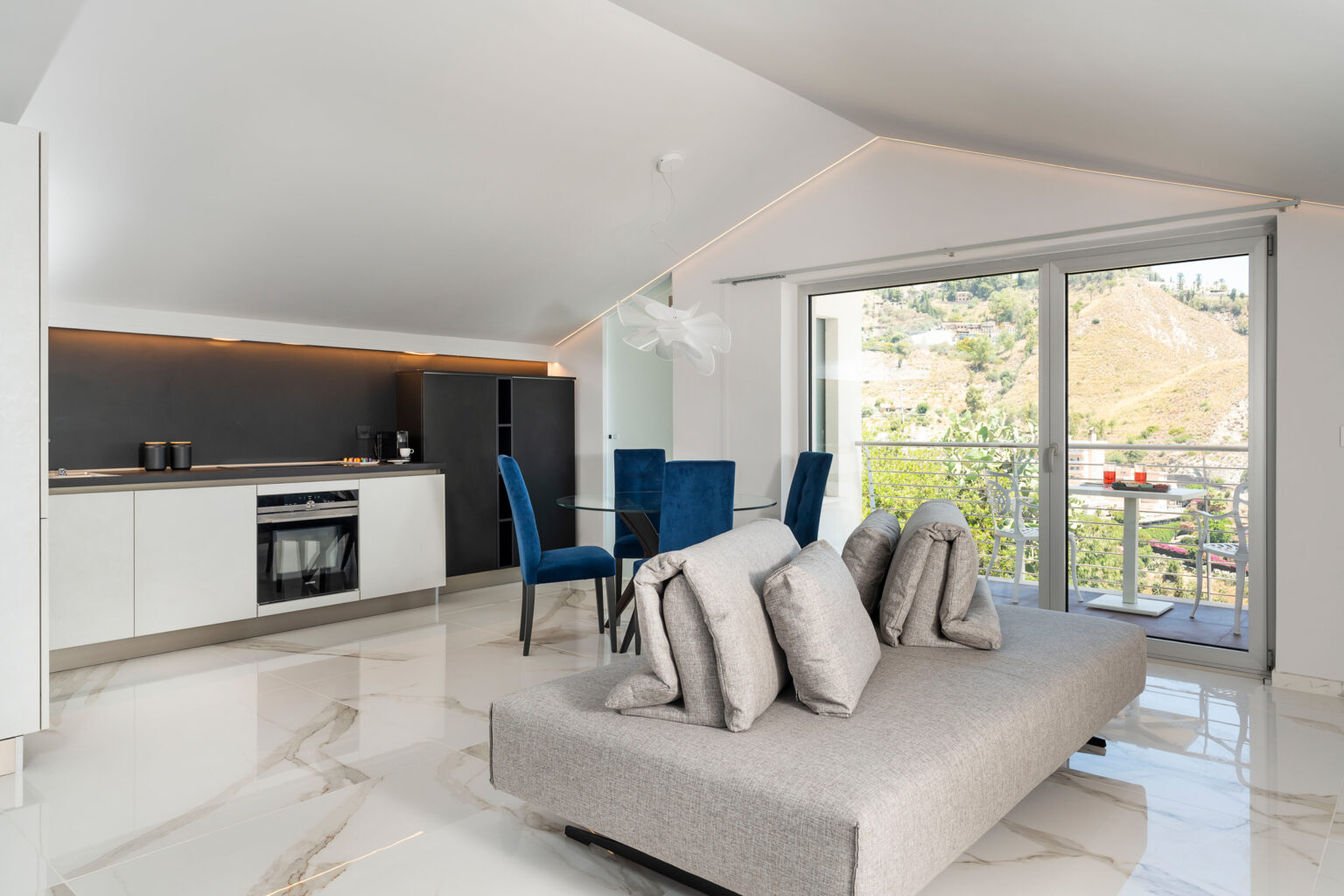 Modern open-plan apartment featuring a sleek kitchen with dark cabinetry, a cozy gray sofa with decorative pillows, and a dining area with blue chairs. Large windows provide natural light and offer a view of the surrounding hills. The polished marble floor adds elegance to the space. | Only on SopranoVillas
