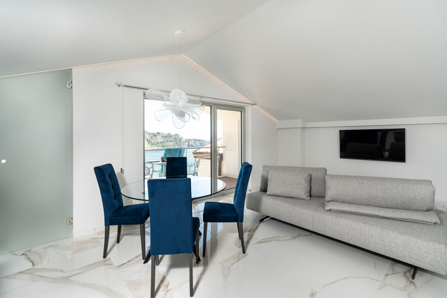 Modern living room with a gray sofa and a glass dining table surrounded by blue chairs, featuring large windows that offer a view of the outdoors. Elegant interior design with white walls and marble flooring. | Only on SopranoVillas