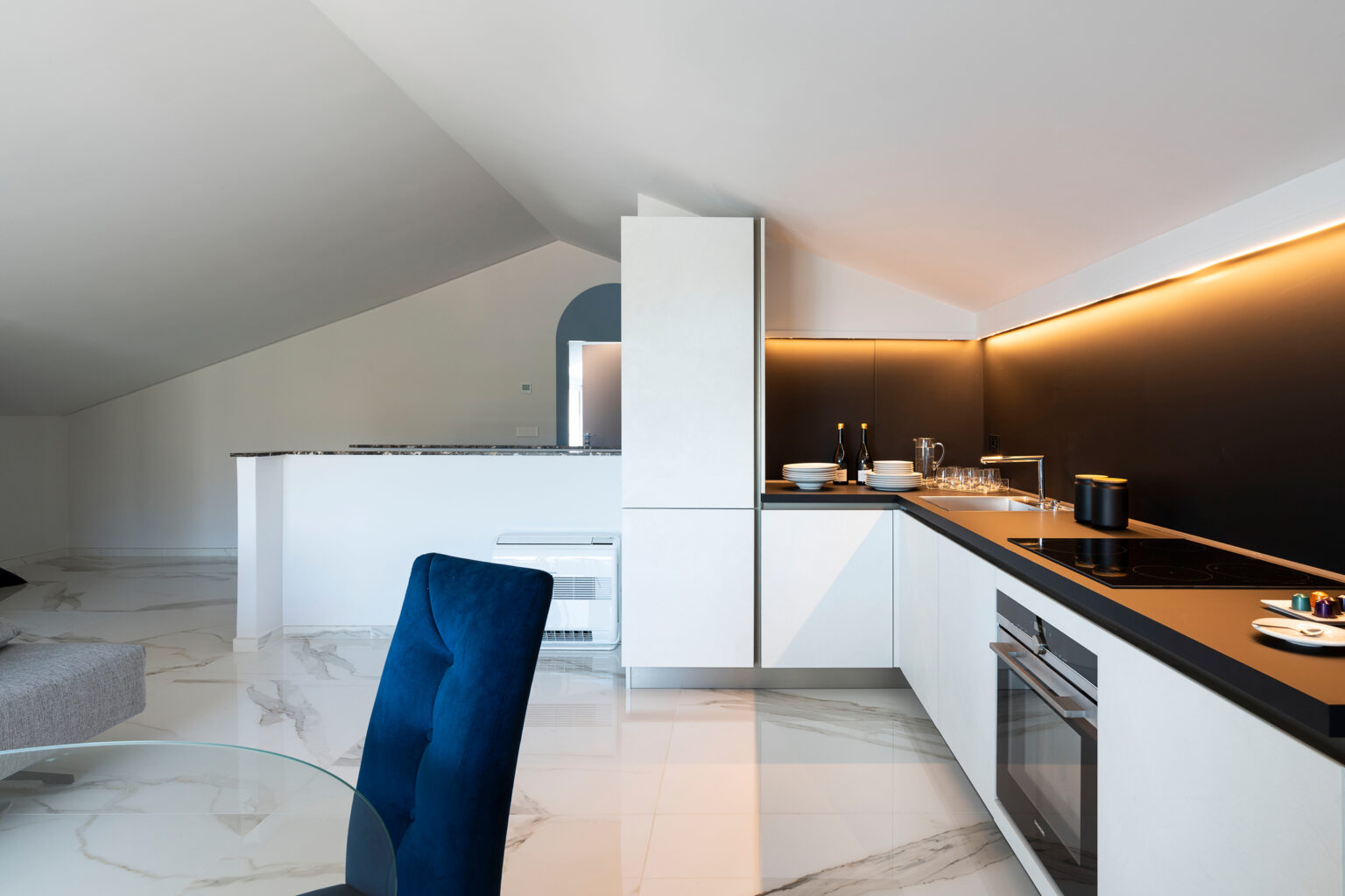 Modern kitchen design featuring sleek white cabinetry, black countertops, and ambient lighting, complemented by a stylish blue chair, in a spacious, bright room with a sloped ceiling. | Only on SopranoVillas