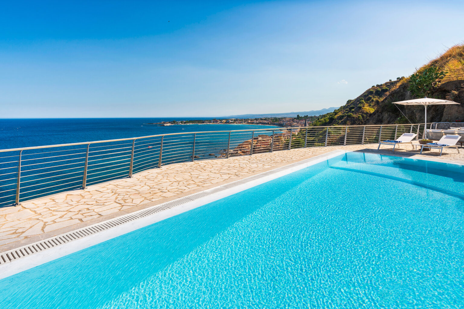 Luxury infinity pool overlooking the ocean with a clear blue sky, surrounded by a scenic coastal landscape and lounge chairs for relaxation. | Only on SopranoVillas