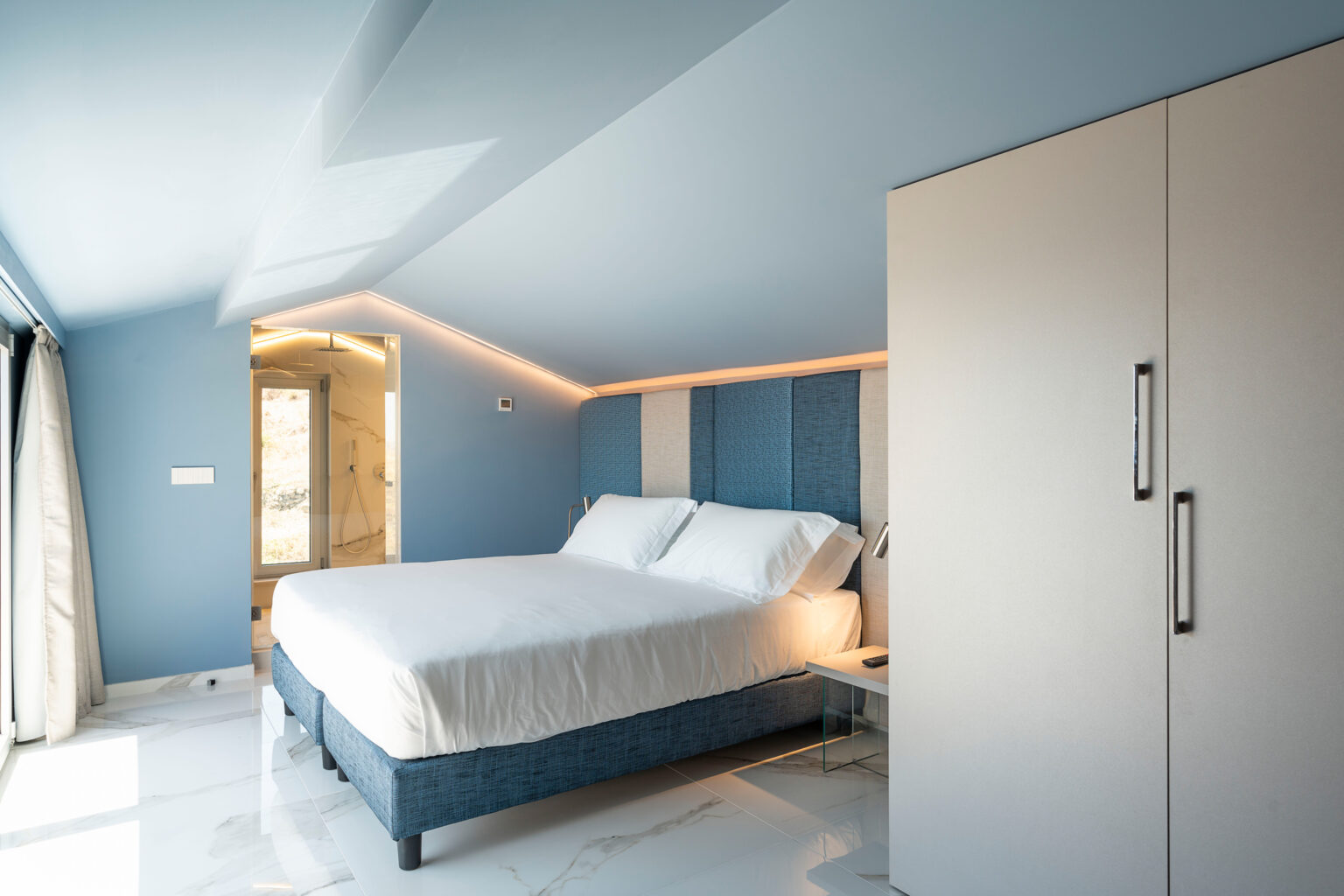 Modern bedroom with a blue accent wall, featuring a queen-sized bed with white linens, a stylish bedside table, and a sleek wardrobe. The room includes a glass-enclosed shower area visible in the background, creating a contemporary and spacious ambiance. Natural light floods the space through large windows, enhancing the airy feel. | Only on SopranoVillas