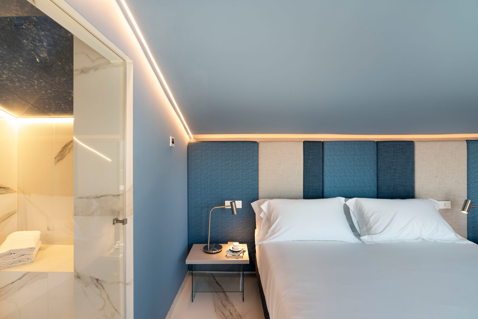 Modern bedroom with a blue accent wall, featuring a cozy bed with white linens, a bedside table with a lamp, and an open bathroom door revealing marble finishes. Soft lighting enhances the contemporary design. | Only on SopranoVillas