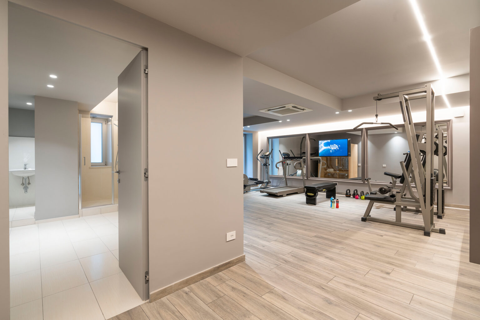 Modern home gym interior with exercise equipment, including weights and a multi-gym setup, featuring sleek design and ample lighting. The space connects to a doorway leading to a bathroom area. | Only on SopranoVillas