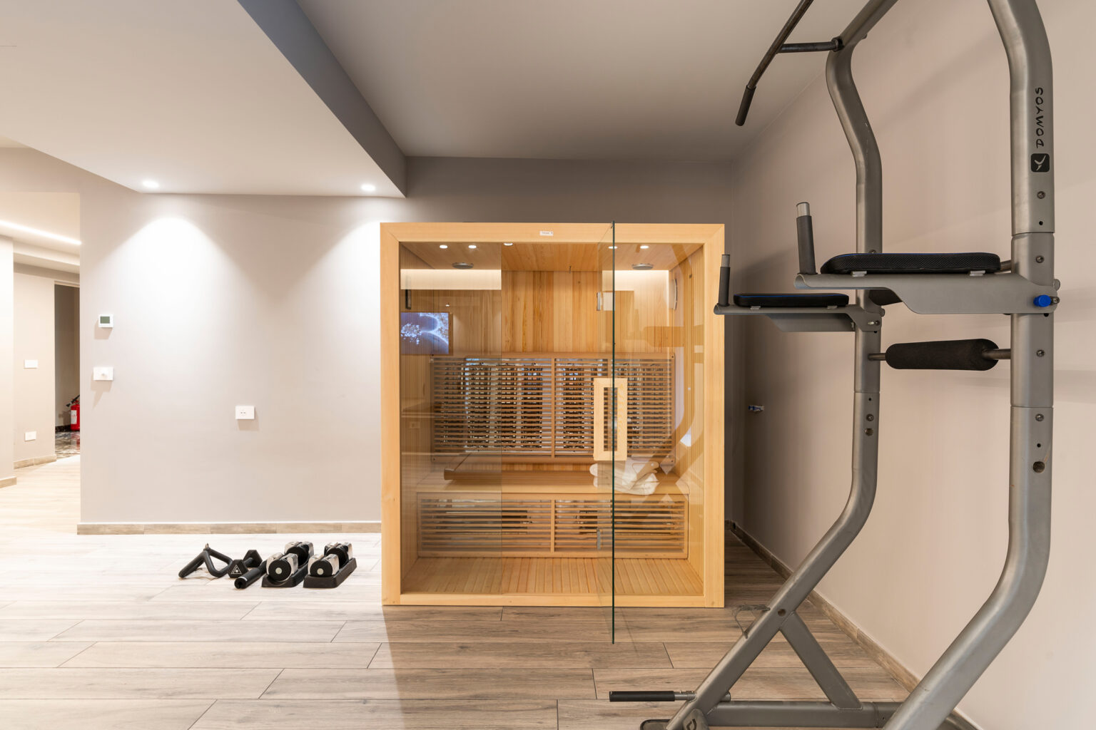 Home gym interior featuring a wooden sauna, a pull-up bar, and dumbbells on the floor, designed for fitness and relaxation. | Only on SopranoVillas
