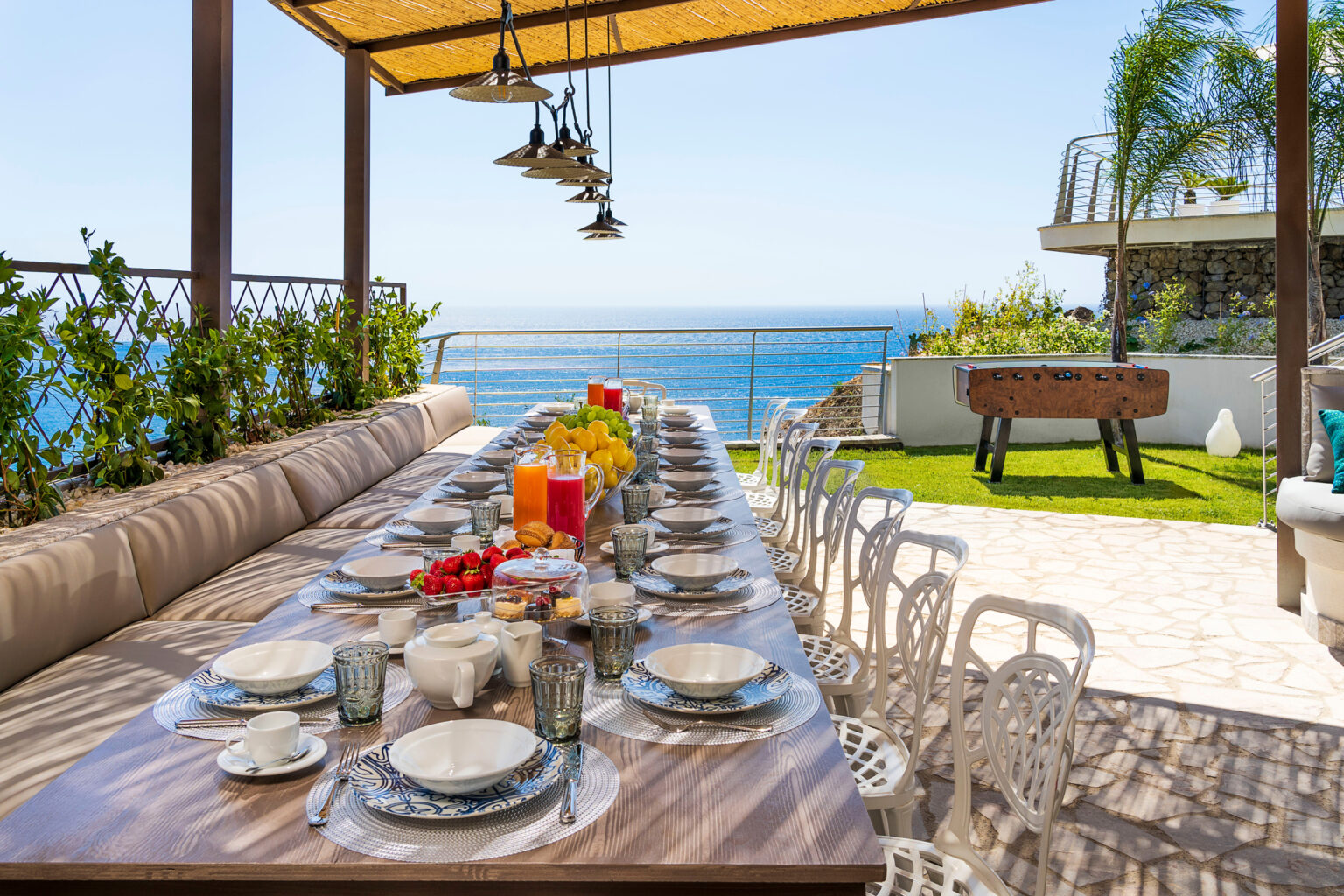 Elegant outdoor dining setup overlooking the ocean, featuring a long wooden table adorned with plates, glasses, and a variety of fresh fruits and beverages, surrounded by lush greenery and a sunny sky. | Only on SopranoVillas