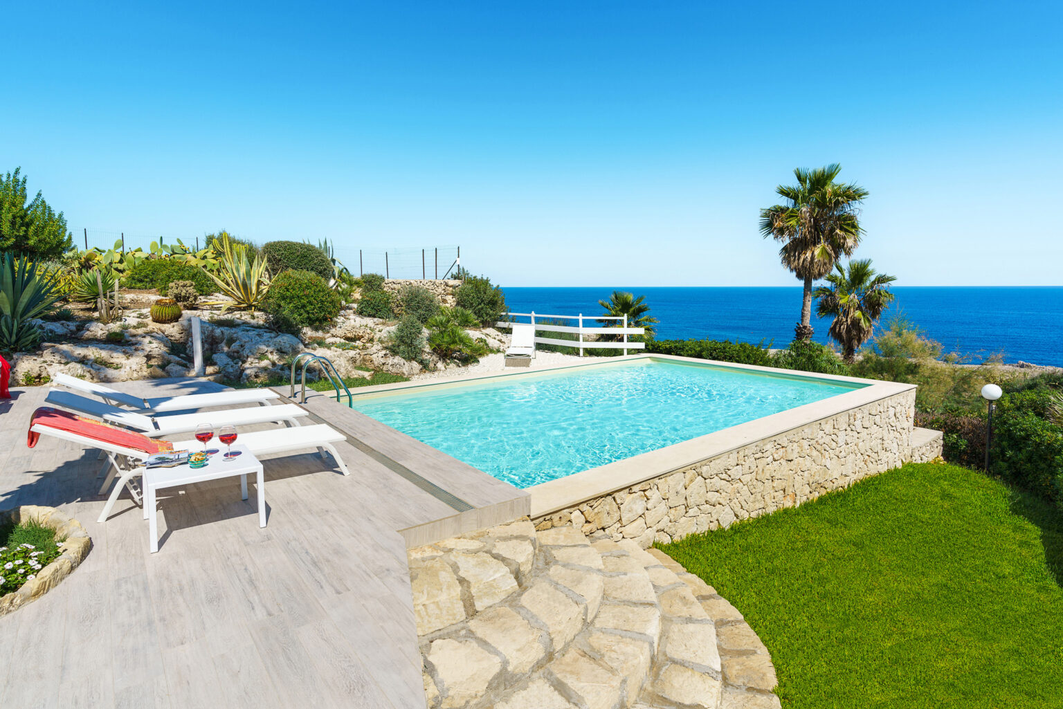 Scenic outdoor pool area featuring a turquoise swimming pool, surrounded by lush greenery and palm trees, with ocean views under a clear blue sky. Two white lounge chairs with a red towel and a small table holding two glasses of red drinks. Perfect for relaxation and leisure. | Only on SopranoVillas
