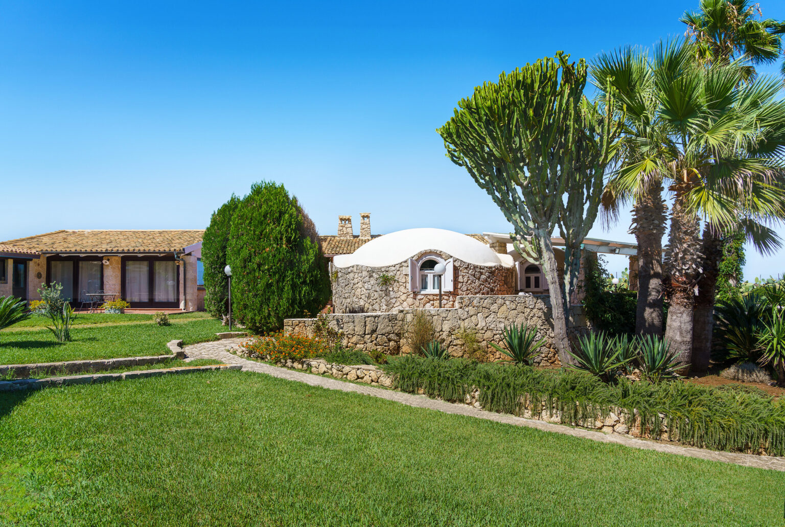 Charming stone cottage surrounded by lush greenery and palm trees, with a clear blue sky above, showcasing a serene outdoor landscape ideal for relaxation and vacation getaways. | Only on SopranoVillas
