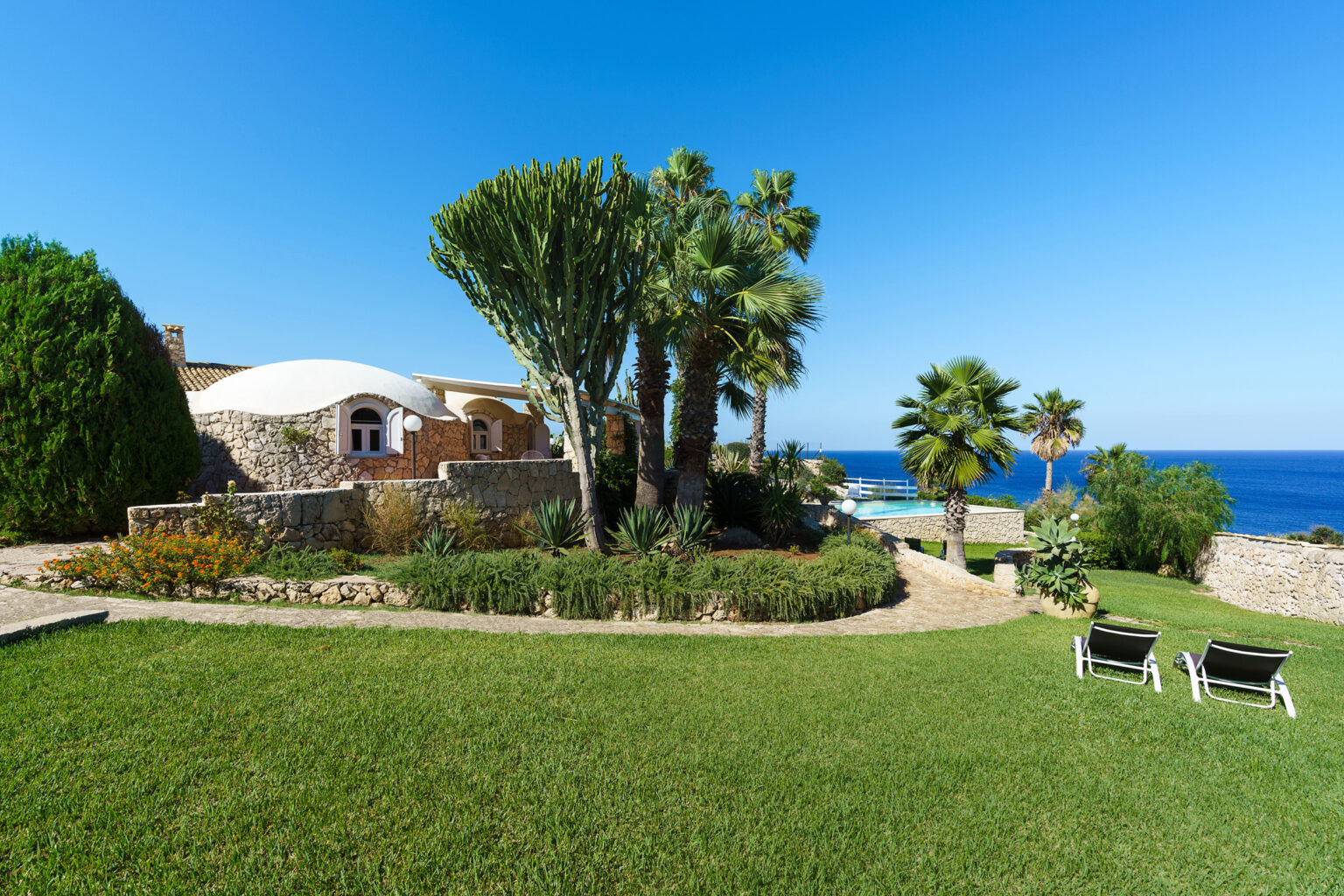 Scenic coastal villa with lush greenery, palm trees, and a well-maintained garden overlooking the ocean, featuring comfortable lounge chairs for relaxation. | Only on SopranoVillas