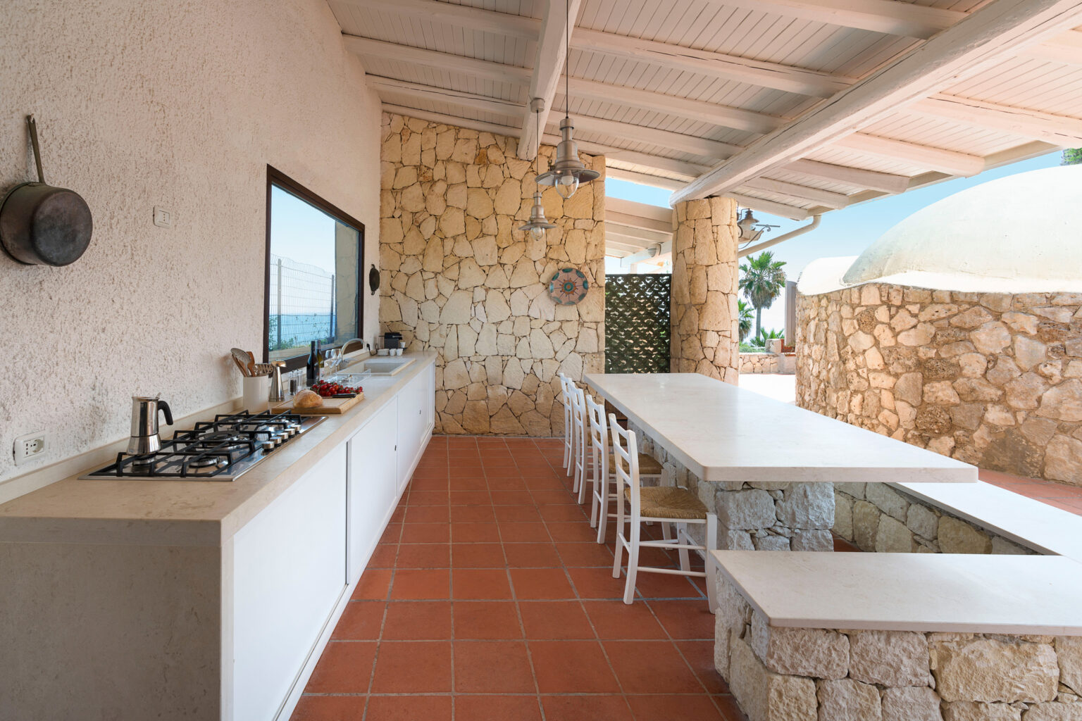 Modern outdoor kitchen with a spacious layout, featuring a gas stove, white cabinetry, and a large dining table, surrounded by natural stone walls and a scenic view. | Only on SopranoVillas
