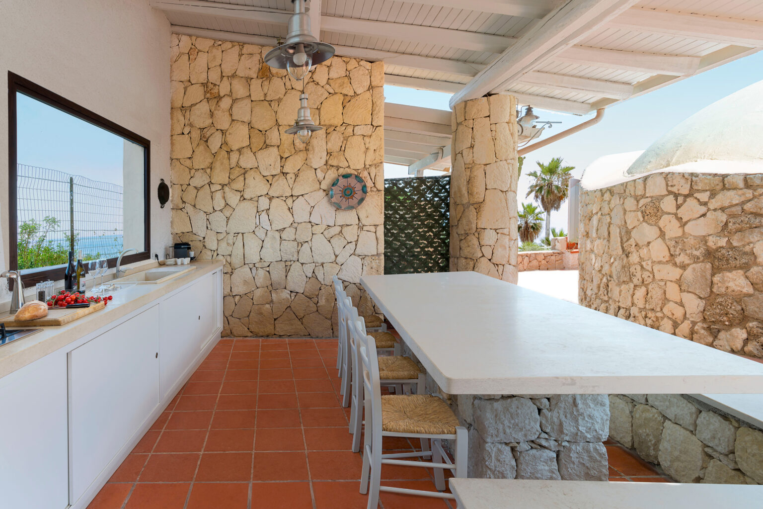 Spacious outdoor kitchen featuring a large stone dining table, white cabinetry, and a stone wall backdrop, with natural light streaming in and views of greenery. Ideal for entertaining and alfresco dining. | Only on SopranoVillas