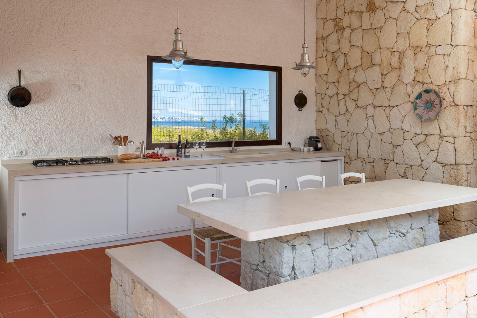 Modern kitchen with stone walls, featuring a large island, white cabinetry, and a scenic window view of vineyards. The space is illuminated by pendant lights, creating a bright and inviting atmosphere. | Only on SopranoVillas