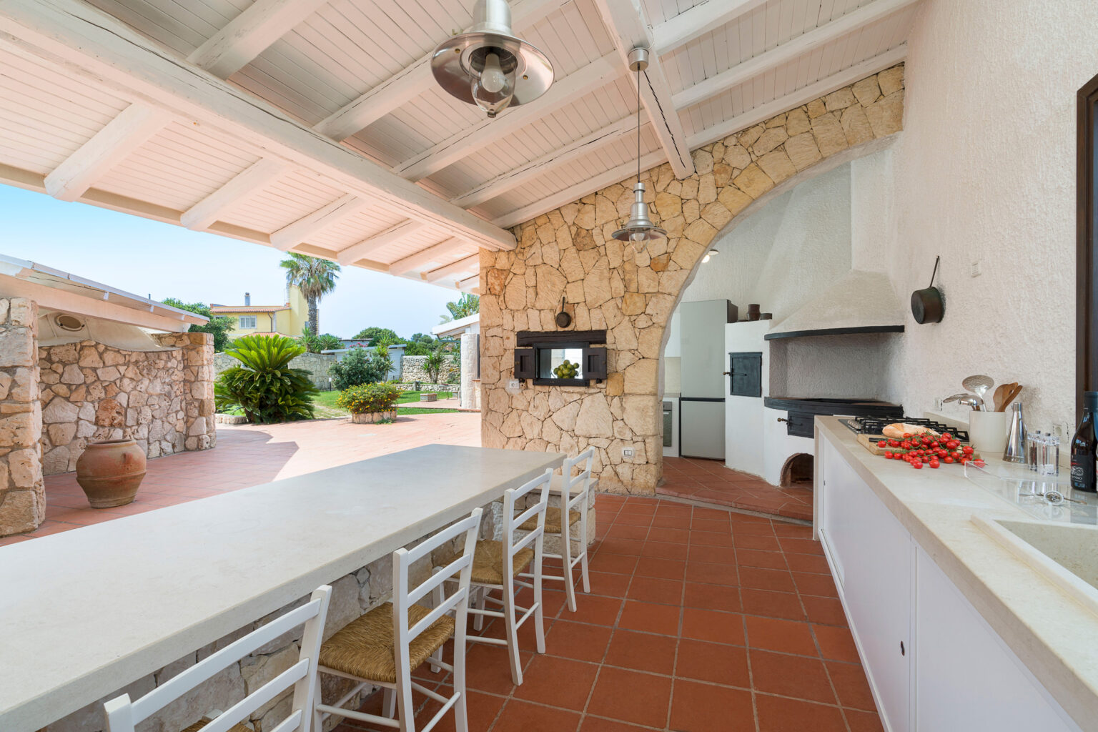 Spacious outdoor kitchen featuring a stone wall, large dining table, and modern appliances, surrounded by greenery and natural light. Ideal for entertaining and al fresco dining. | Only on SopranoVillas