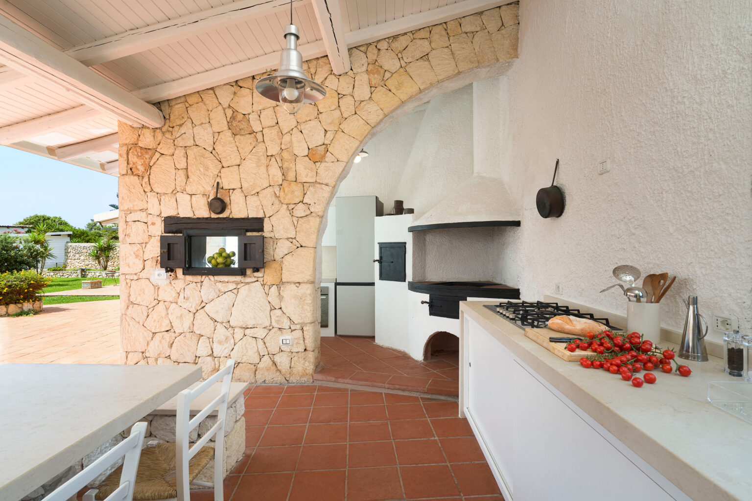 Modern outdoor kitchen with stone archway, featuring a barbecue grill, white cabinetry, and fresh tomatoes on the counter, set in a bright, inviting space. | Only on SopranoVillas