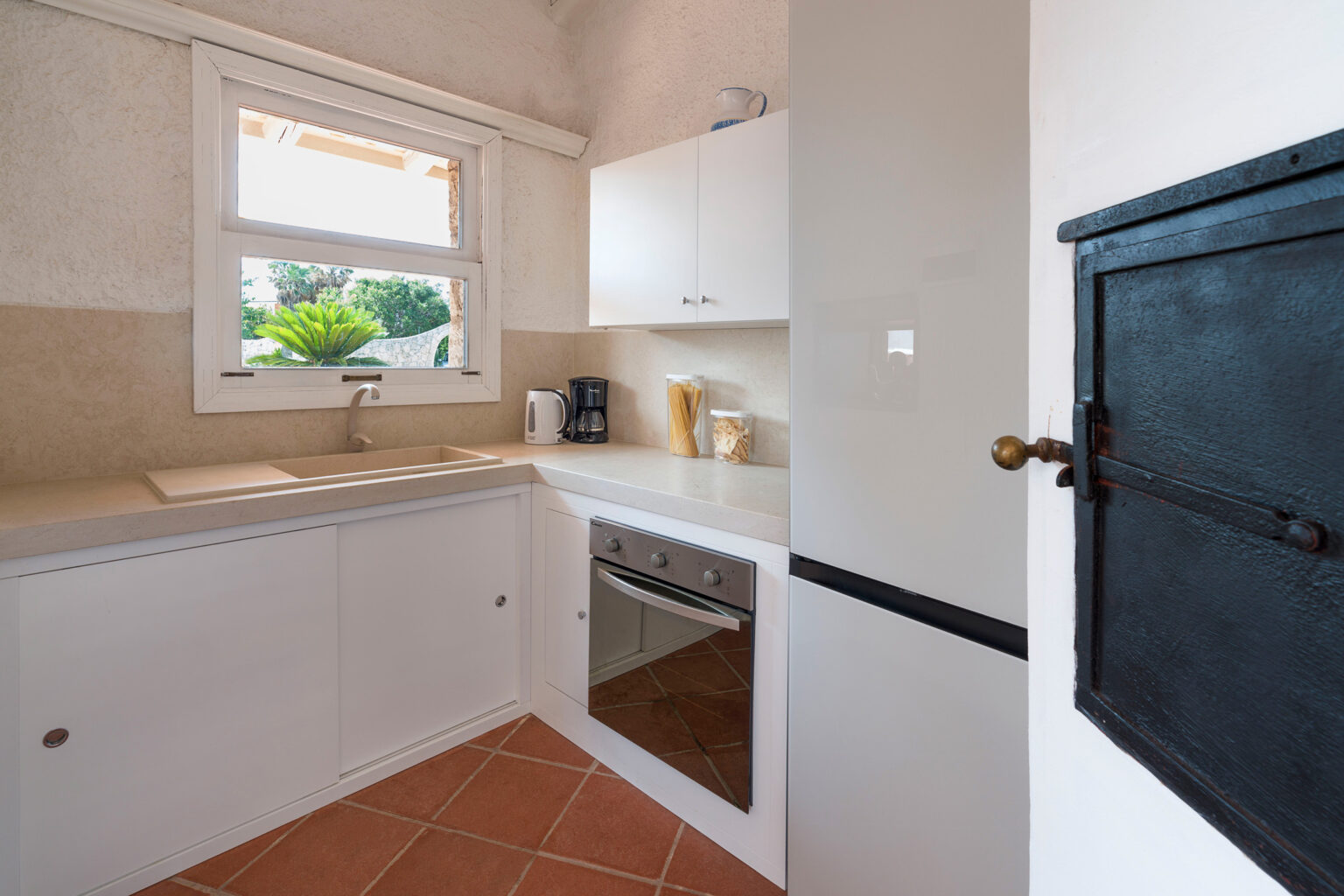 Modern kitchen interior featuring white cabinetry, a built-in oven, and a window with greenery outside, complemented by a rustic black mailbox. | Only on SopranoVillas