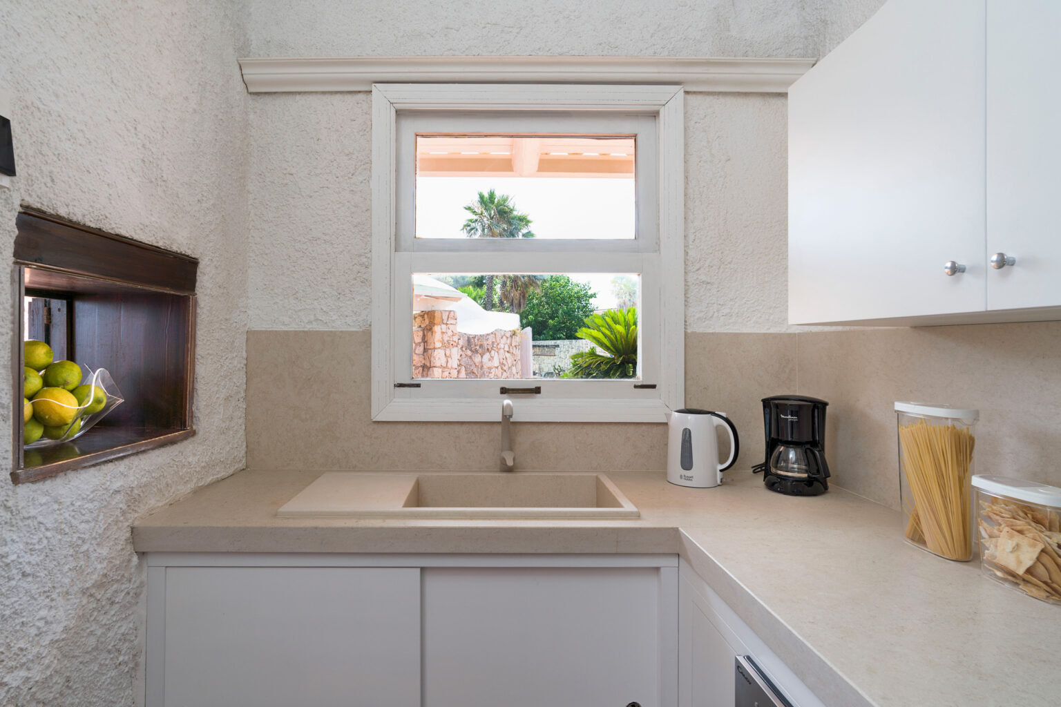 Bright and modern kitchen featuring a sink, coffee maker, and a window with a view of greenery, complemented by light-colored cabinets and a minimalist design. | Only on SopranoVillas