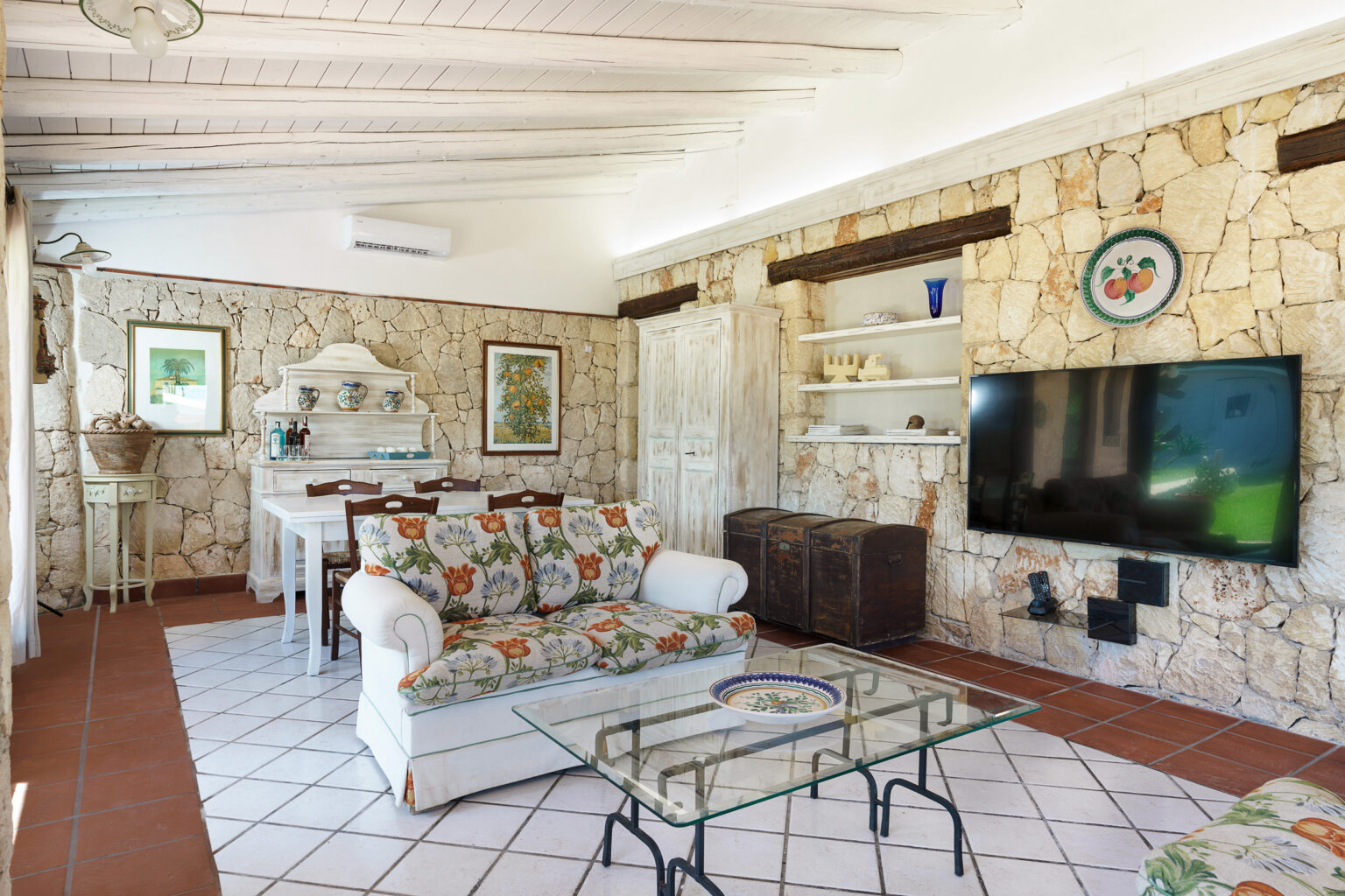Cozy living room with stone walls, featuring a comfortable floral-patterned sofa, glass coffee table, and a large flat-screen TV. The space includes a dining area with white furniture and decorative art on the walls, creating a warm and inviting atmosphere. | Only on SopranoVillas