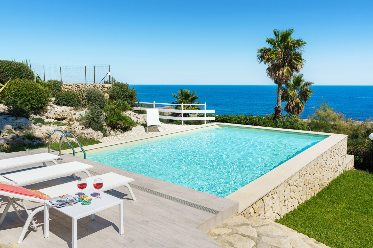 Luxury outdoor pool with stunning ocean view, surrounded by palm trees and lush greenery. Two sun loungers and a table with drinks and snacks create an inviting relaxation space. Perfect for summer getaways and tranquil retreats. | Only on SopranoVillas