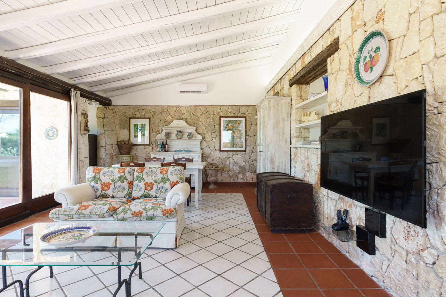 Cozy living room with stone walls, a floral-patterned sofa, and a large flat-screen TV, featuring a glass coffee table and decorative wall art, creating a welcoming atmosphere. | Only on SopranoVillas