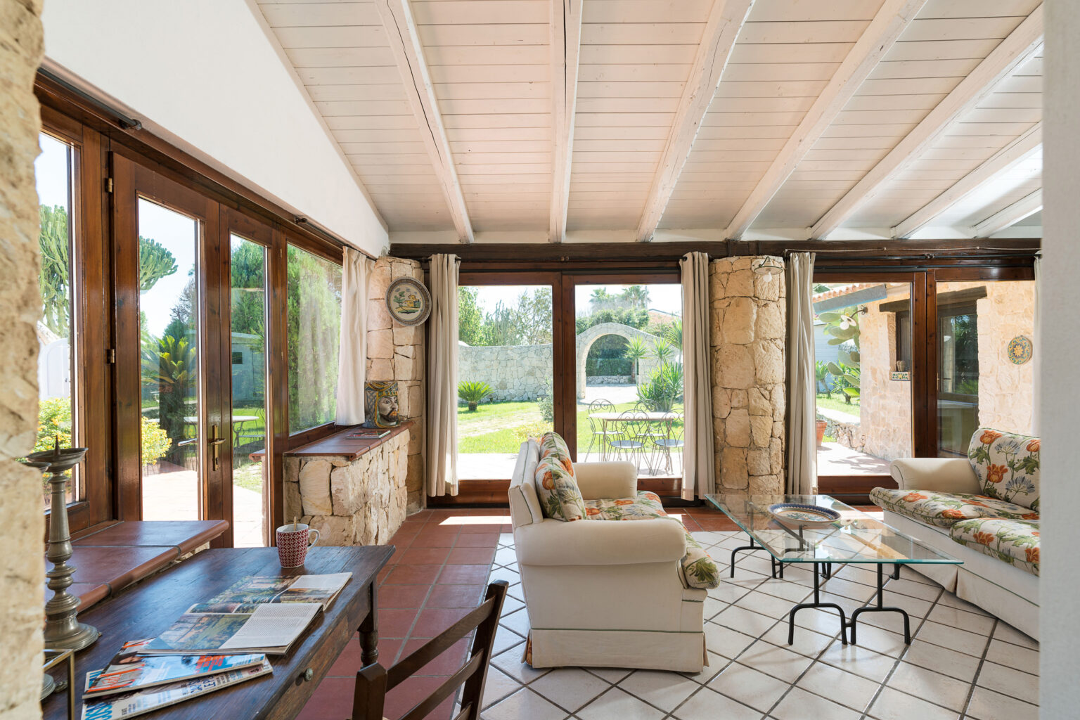 Bright and airy sunroom with large windows, wooden beams, and stone accents, featuring comfortable seating and a view of a lush garden. Ideal for relaxation and indoor-outdoor living. | Only on SopranoVillas