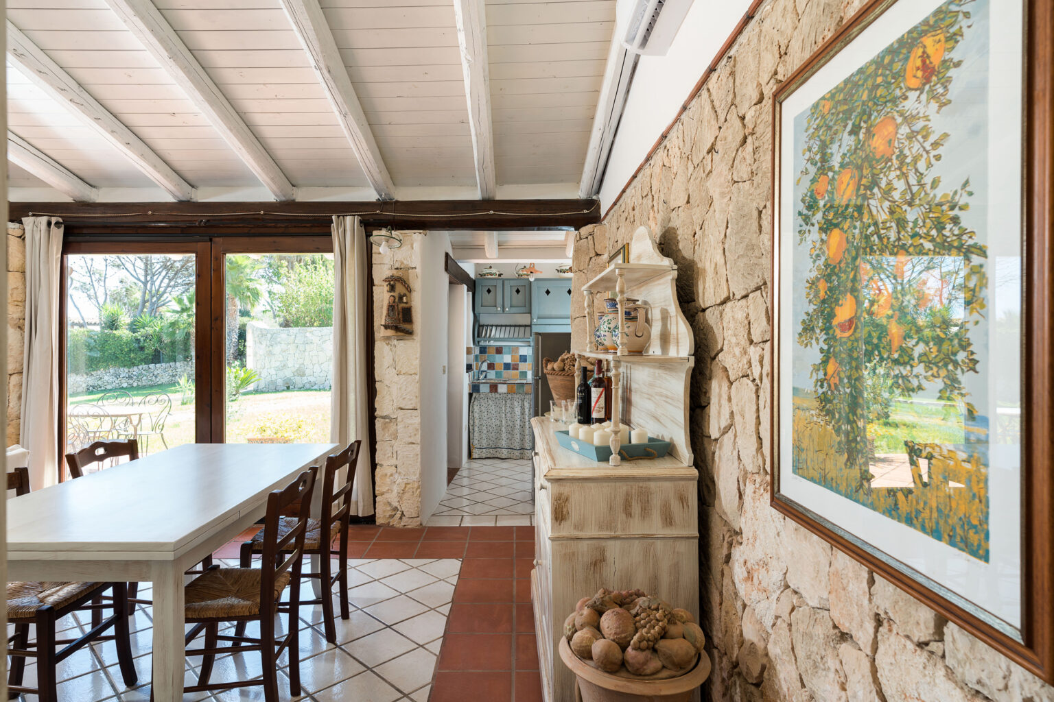 Interior view of a rustic kitchen and dining area featuring a long white table, stone walls, and a vibrant fruit-themed painting, with natural light streaming through large windows that open to a garden. | Only on SopranoVillas